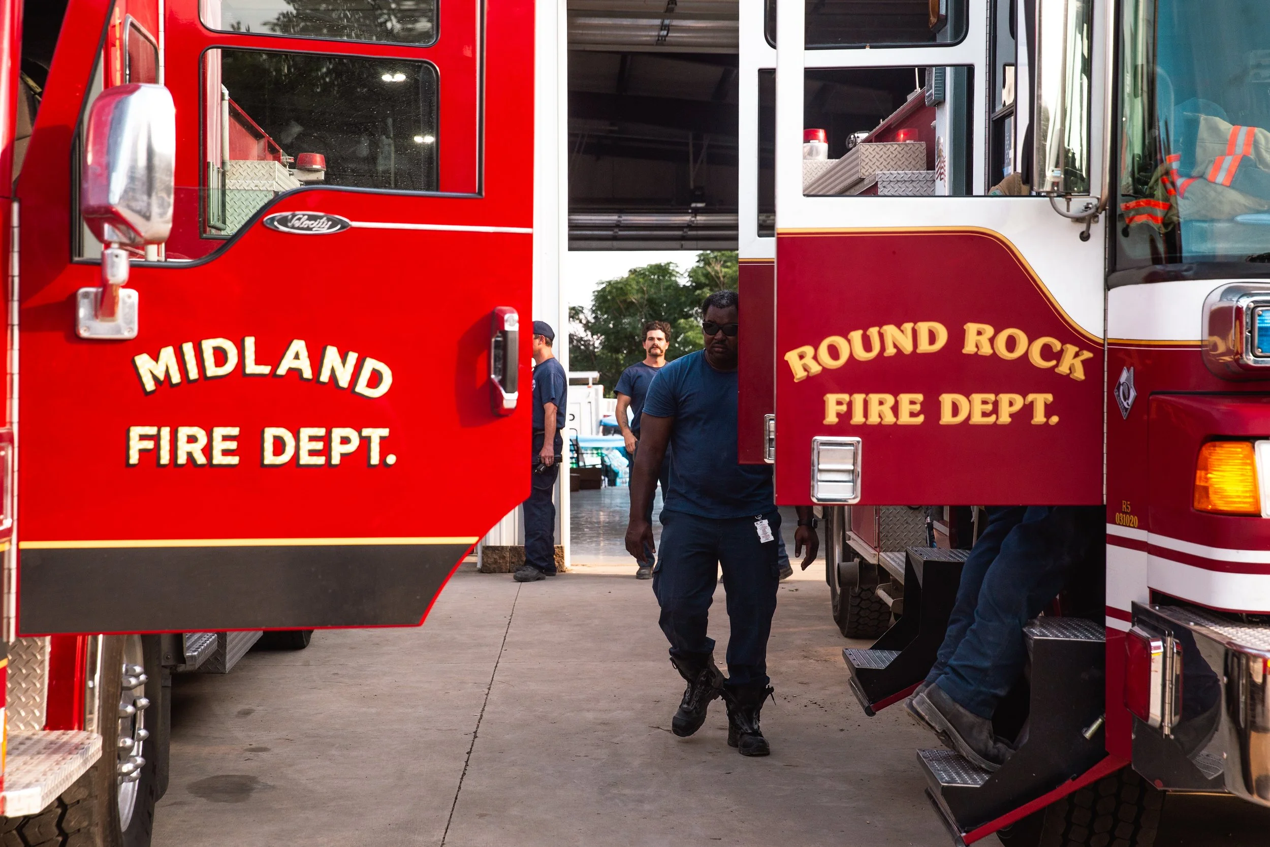 Firefighters in uniform walking between red fire trucks labeled 'Midland Fire Dept.' and 'Round Rock Fire Dept.' in a fire station.