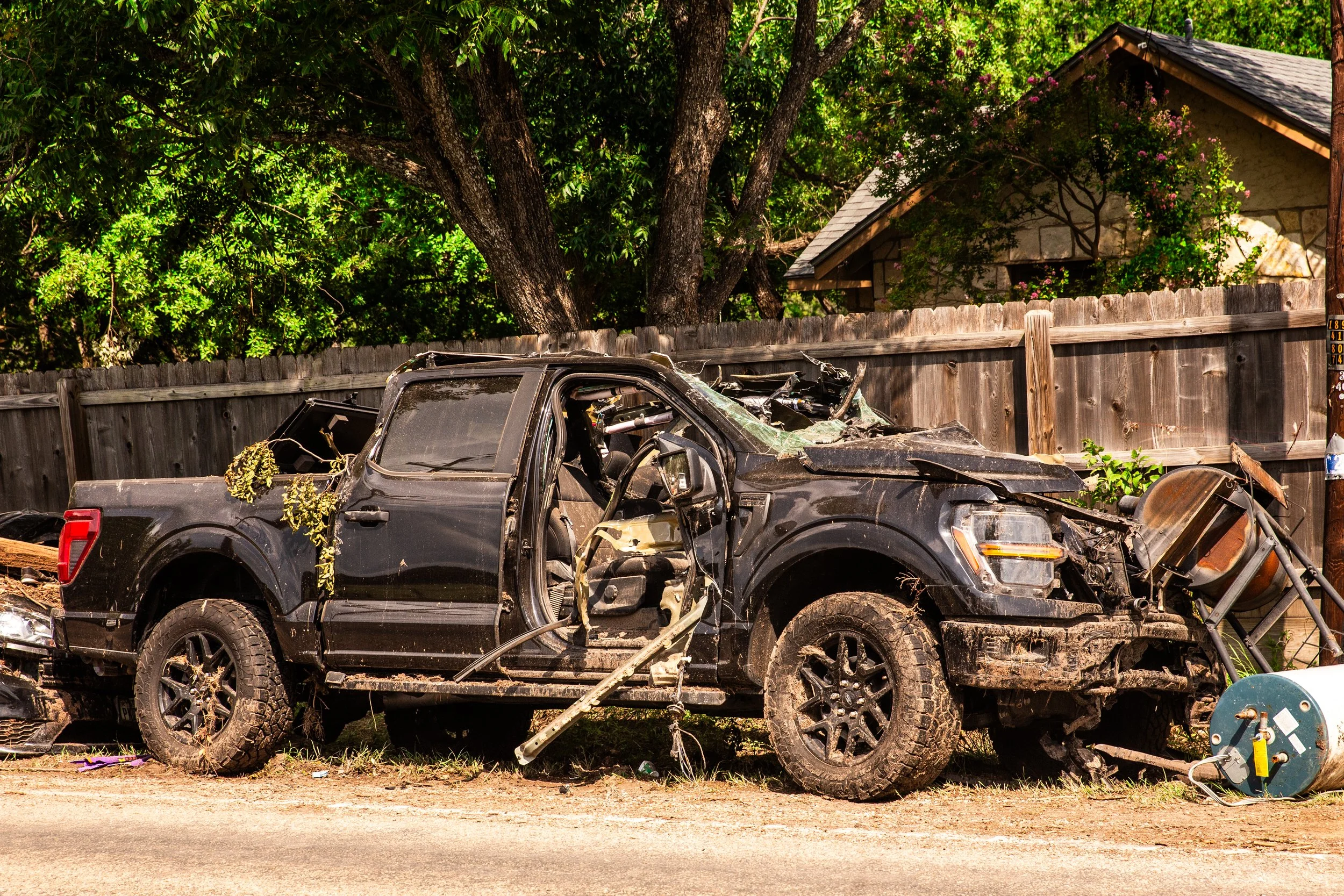 A severely damaged black pickup truck with a crumpled front end and a missing door, parked on the side of a road, with a fallen streetlight pole nearby. The truck is muddy and surrounded by dirt, with a residential backyard fence and trees in the bac
