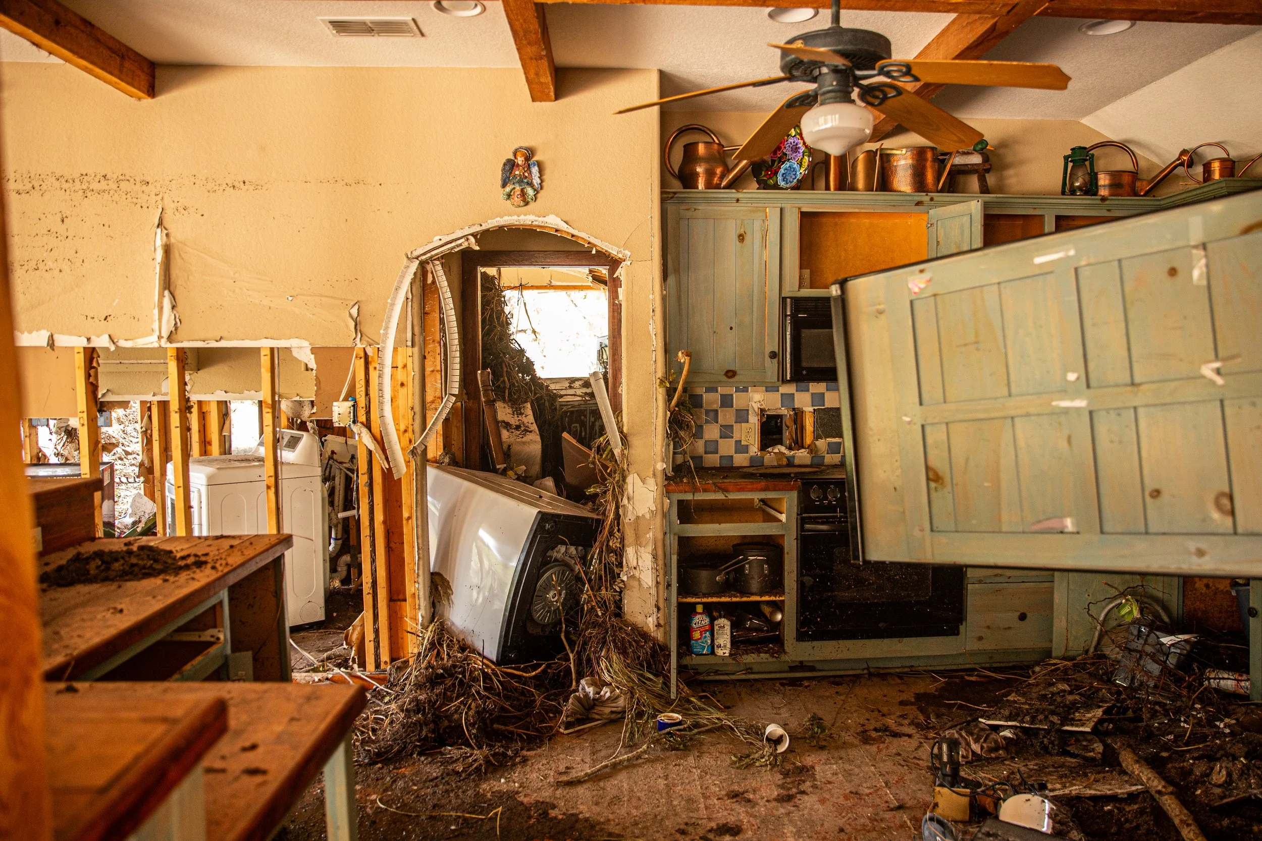 Interior of a damaged kitchen with a collapsed ceiling, broken wall, and scattered debris, including a washing machine, cabinets, and kitchen appliances.