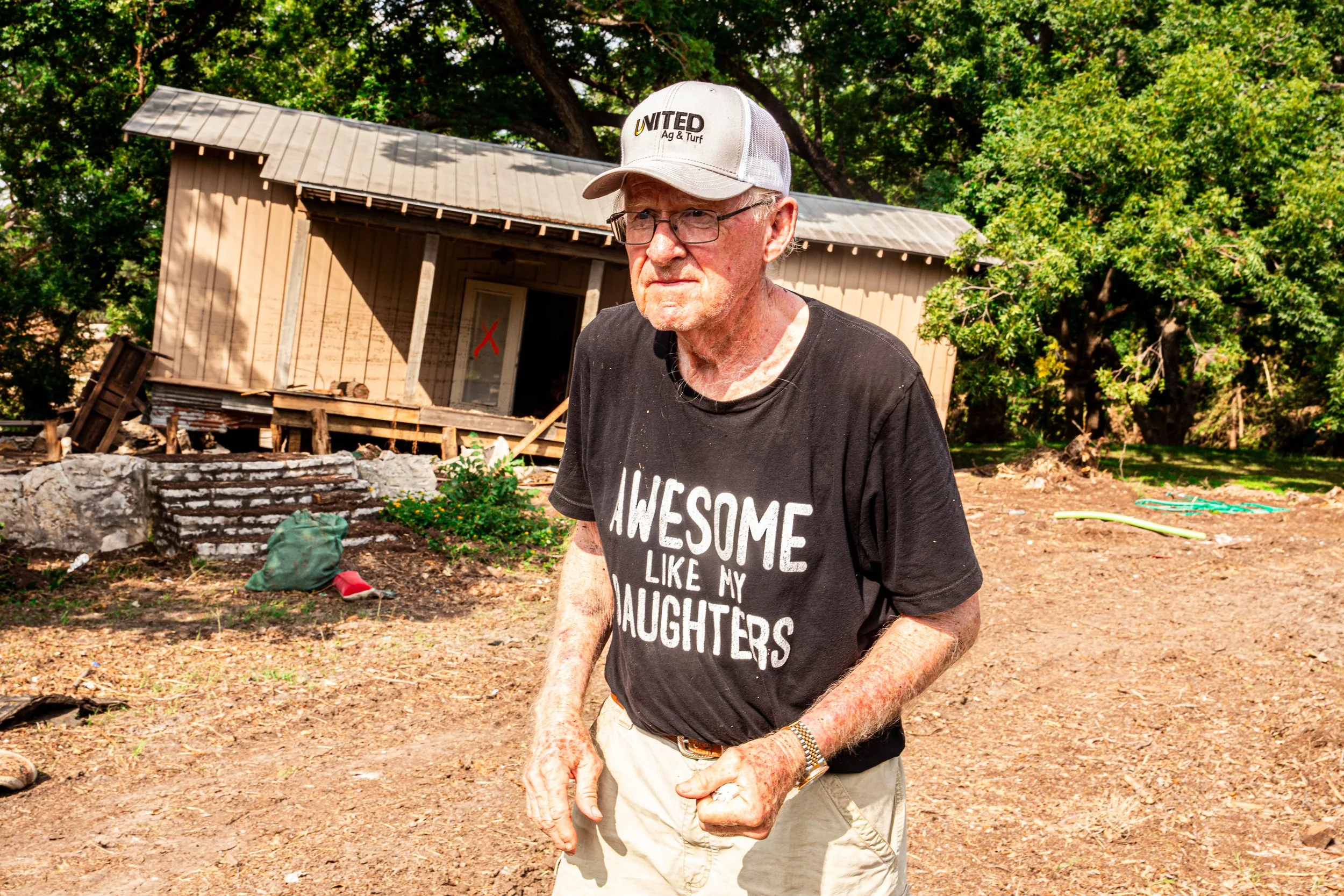 An elderly man wearing a black T-shirt with white text, a baseball cap, and glasses standing outdoors with a partially constructed wooden building and green trees in the background.