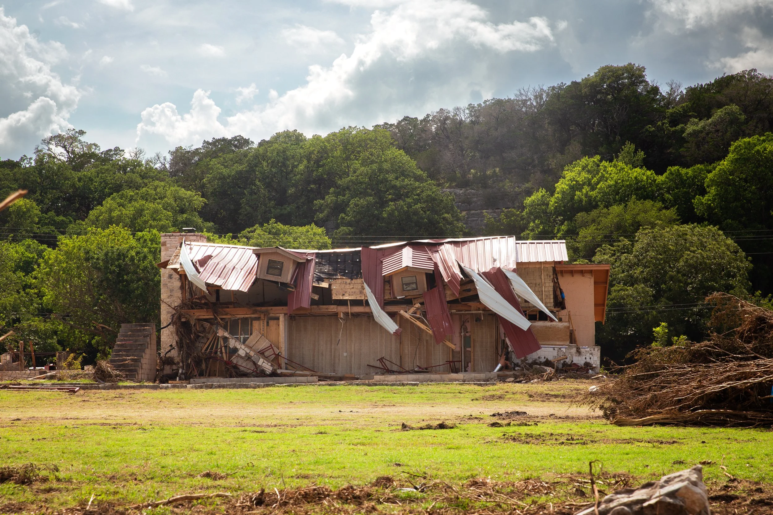A house with significant roof damage, with parts of the roof collapsed and hanging down, set in a green area with trees in the background.