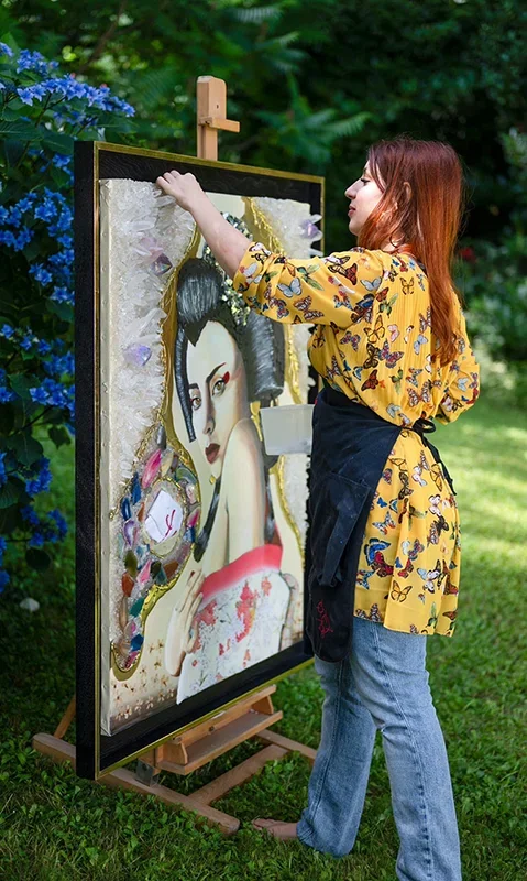 Une femme peint sur une toile dans un jardin, avec des fleurs bleues à gauche.