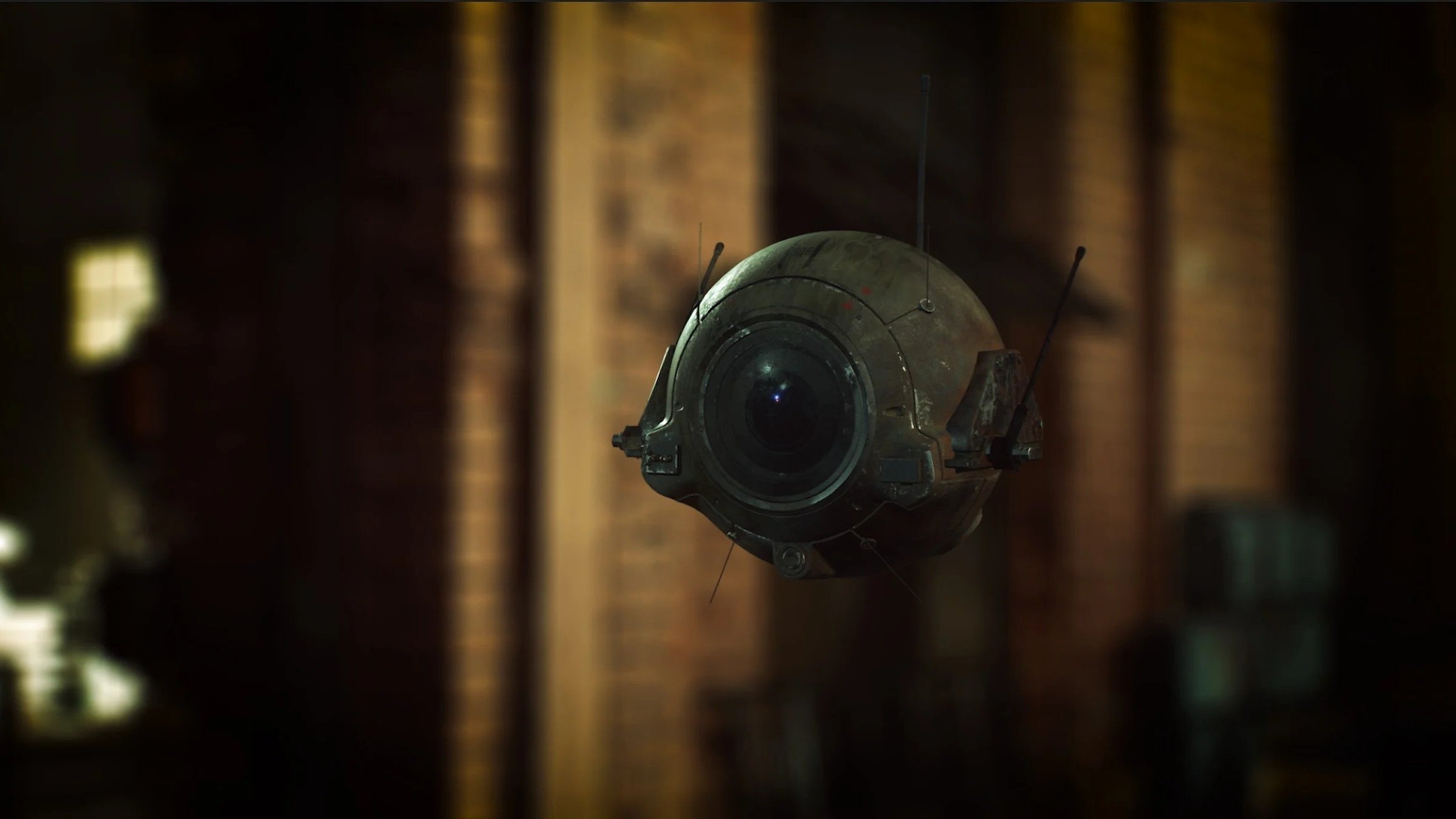 A small, round robot with a large central eye or camera, floating indoors against a blurry background of wooden walls or panels.