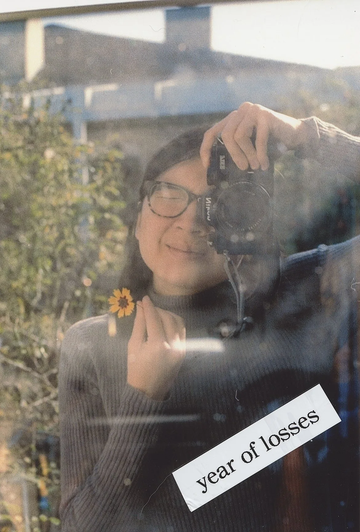A woman taking a mirror selfie outdoors, holding a yellow flower near her chin, with a reflection of trees and buildings in the background.