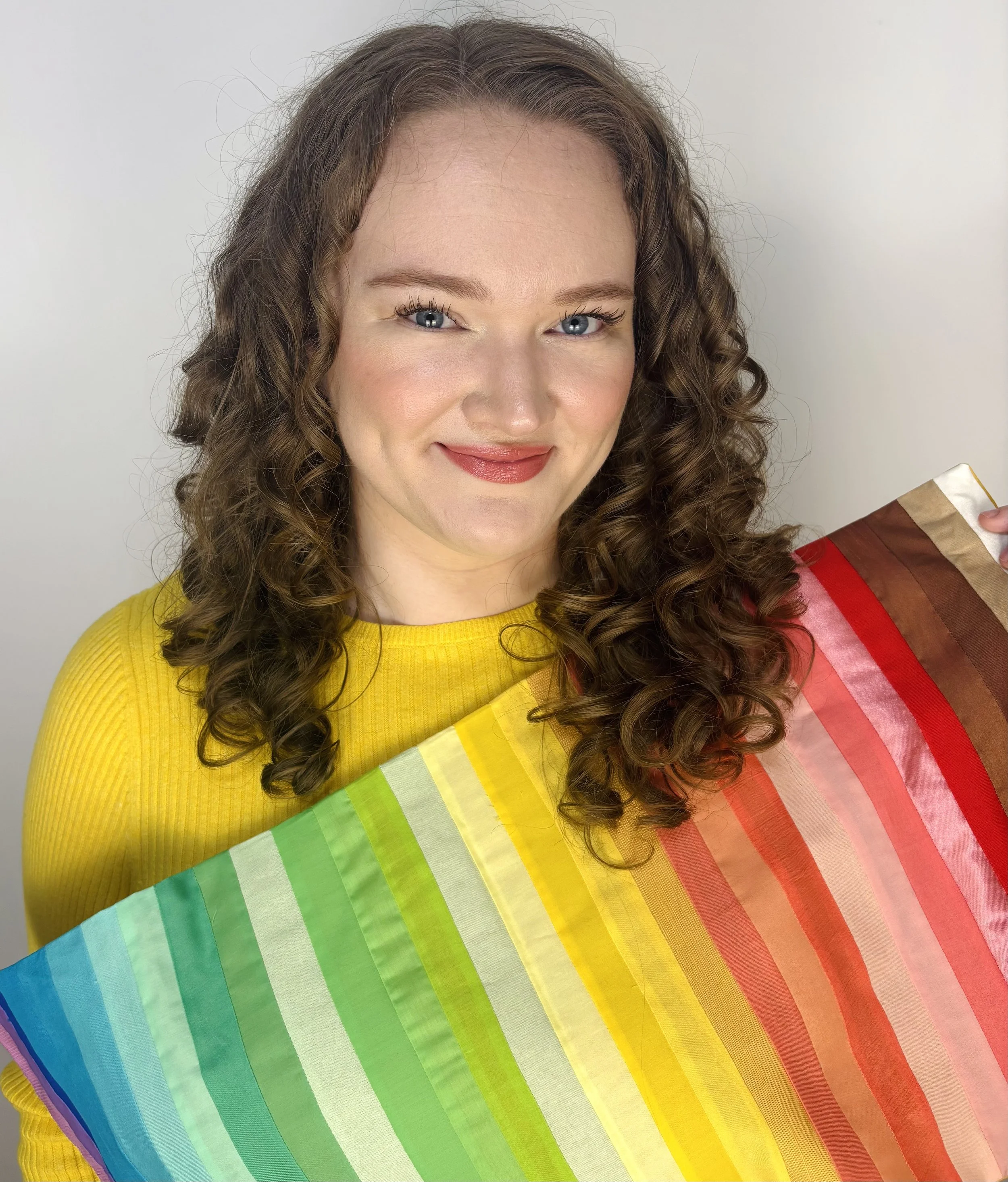 A woman with curly brown hair wearing a yellow sweater, smiling slightly, holding a rainbow-colored fabric with rainbow stripes.