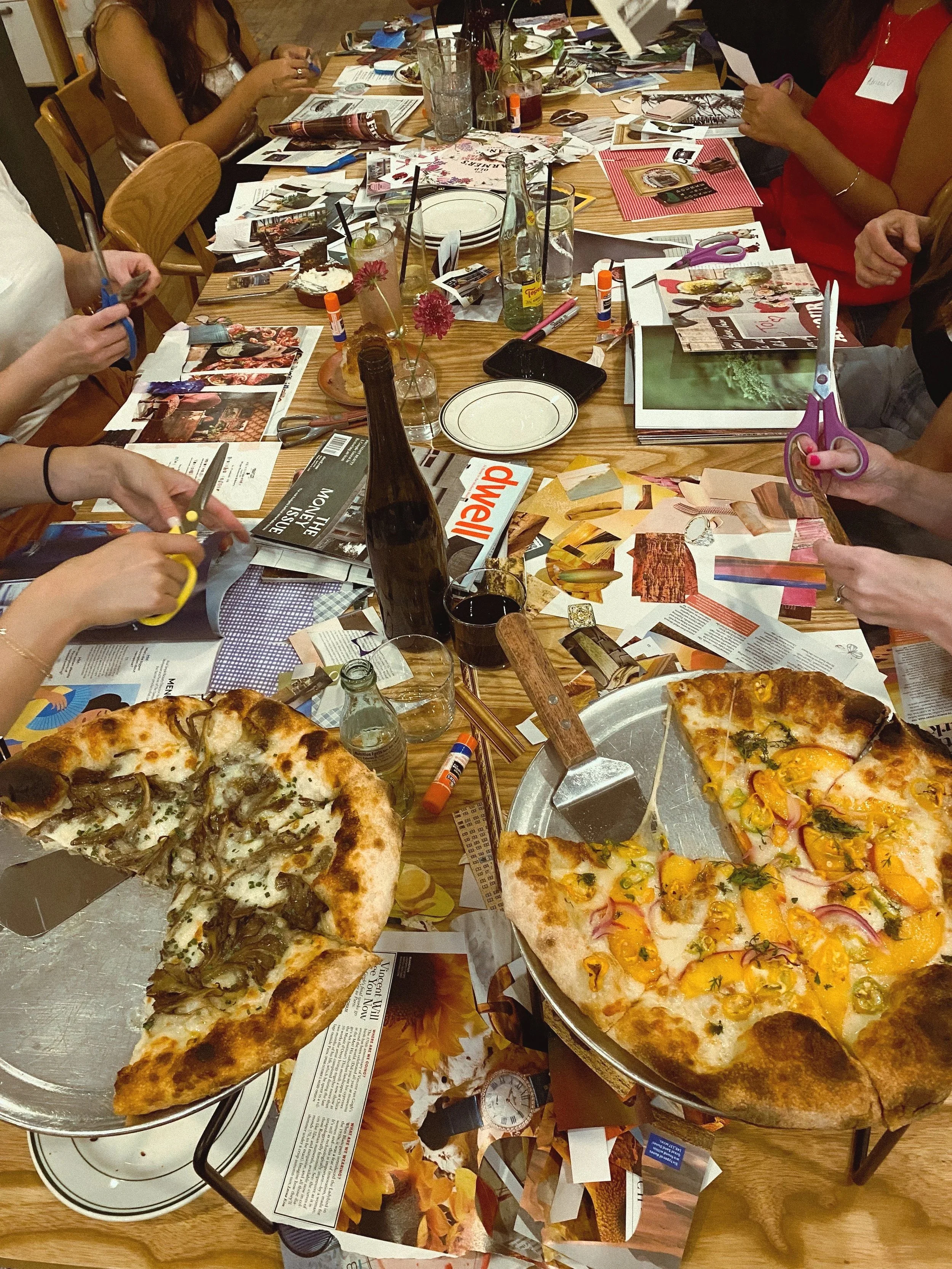 A table with two pizzas, one with mushrooms and the other with peaches and red onion, surrounded by scattered magazines, scissors, glue sticks, glasses, bottles, and people holding scissors and collaging.