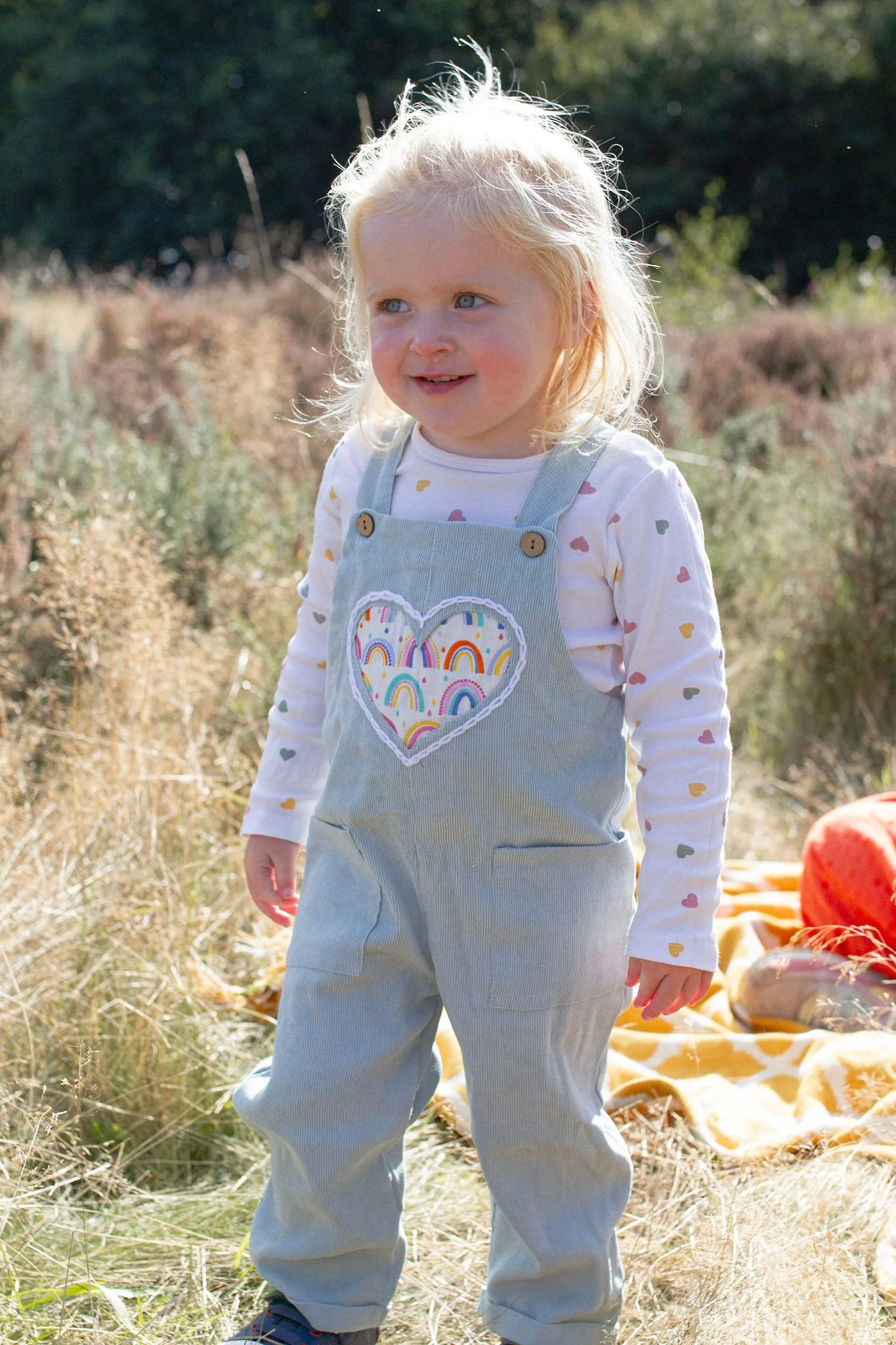 A young girl with blonde hair and blue eyes standing outdoors in a field of dry grass, wearing gray overalls with a colorful rainbow heart patch on the front and a white long-sleeve shirt with multicolored heart patterns.
