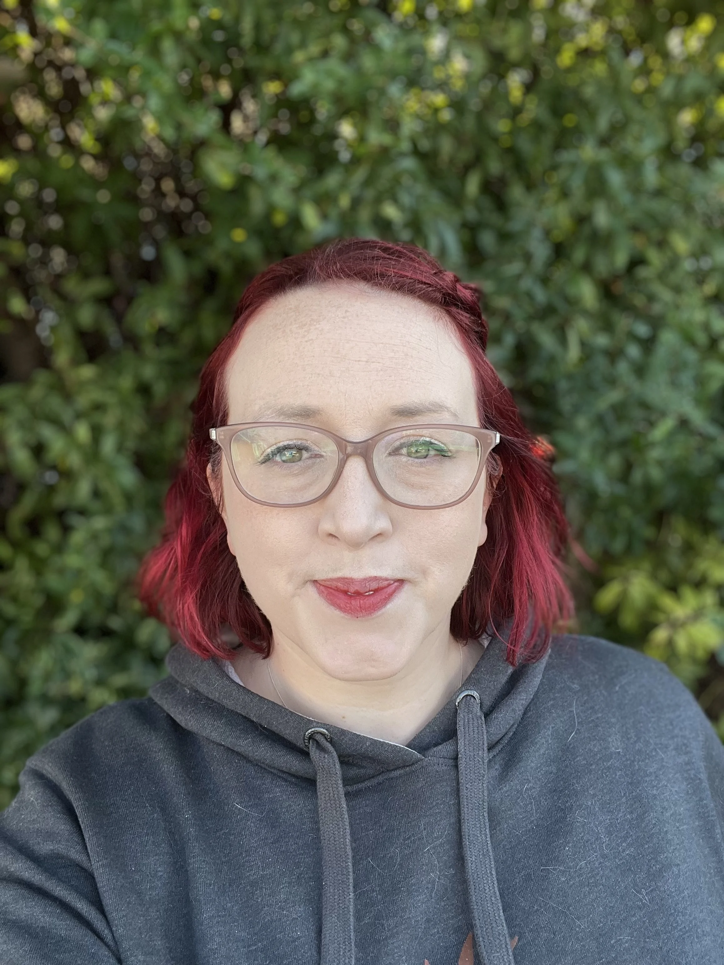 A woman with red hair and glasses taking a selfie outdoors, standing in front of green leafy bushes.