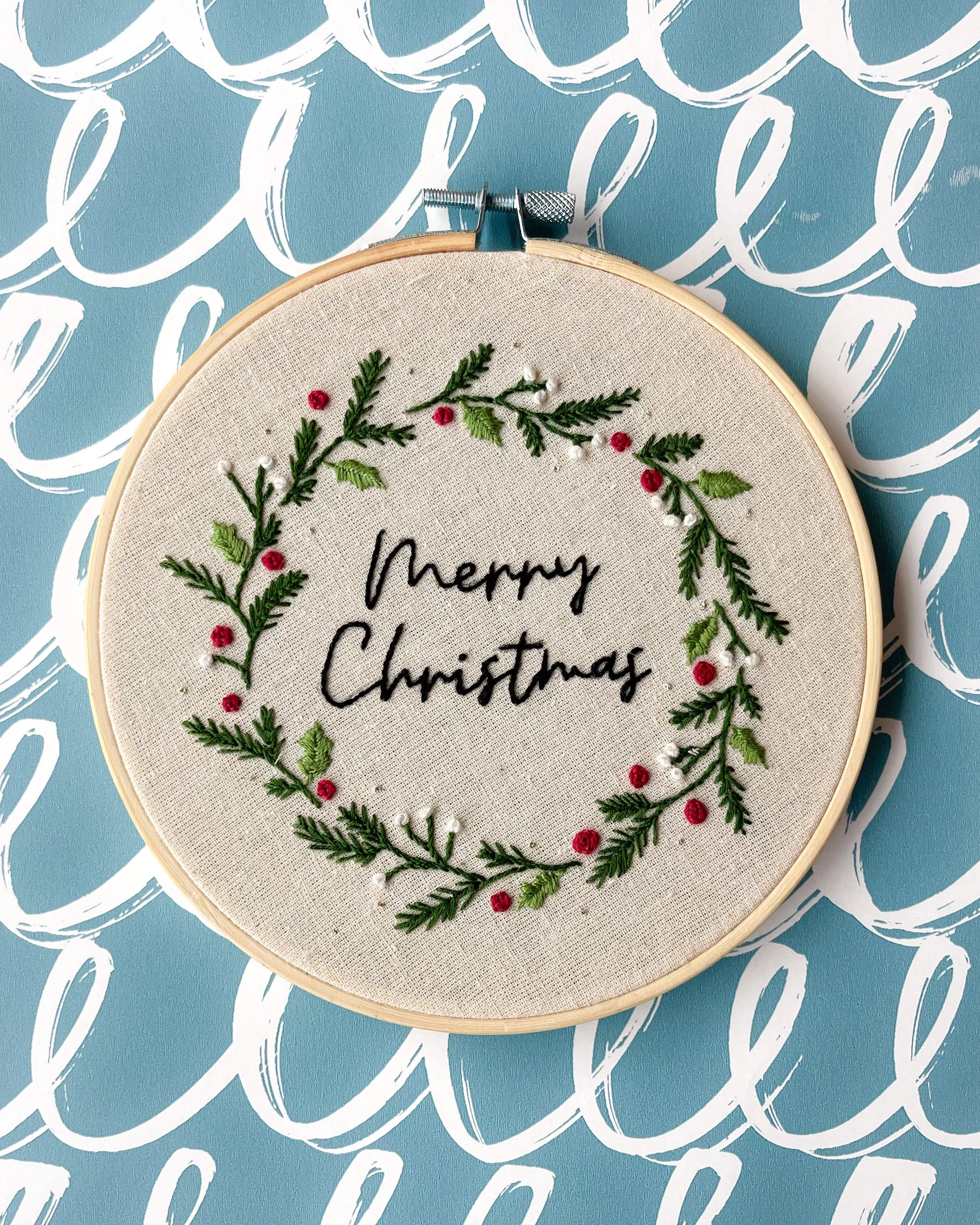 Embroidery hoop with a Christmas wreath design and the words 'Merry Christmas' embroidered in the center, set against a blue background with white cursive writing.