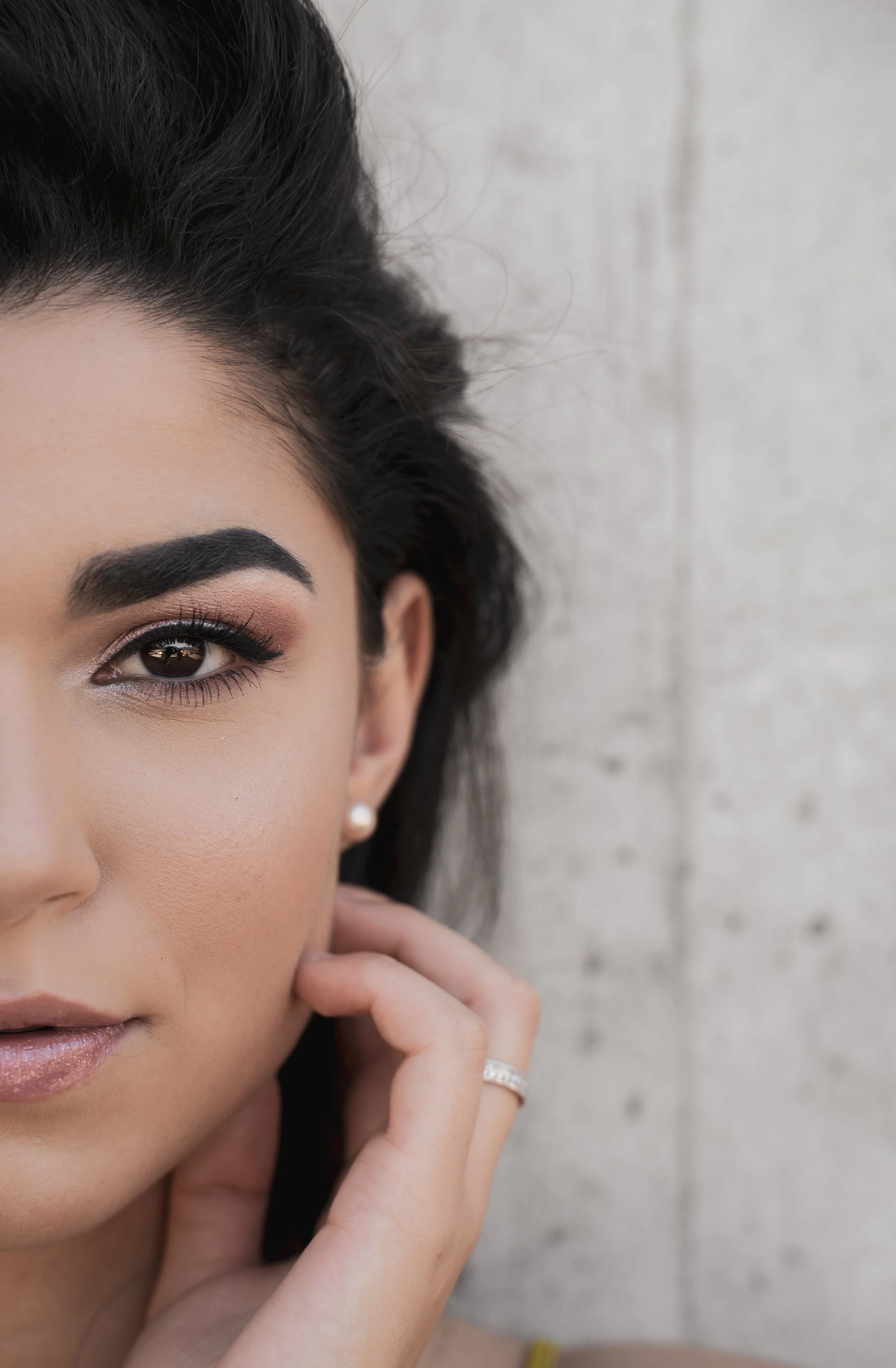 Gros plan d'une femme avec maquillage, portant une boucle d'oreille en perle, posant près d'un mur en pierre, avec une main près de son visage.