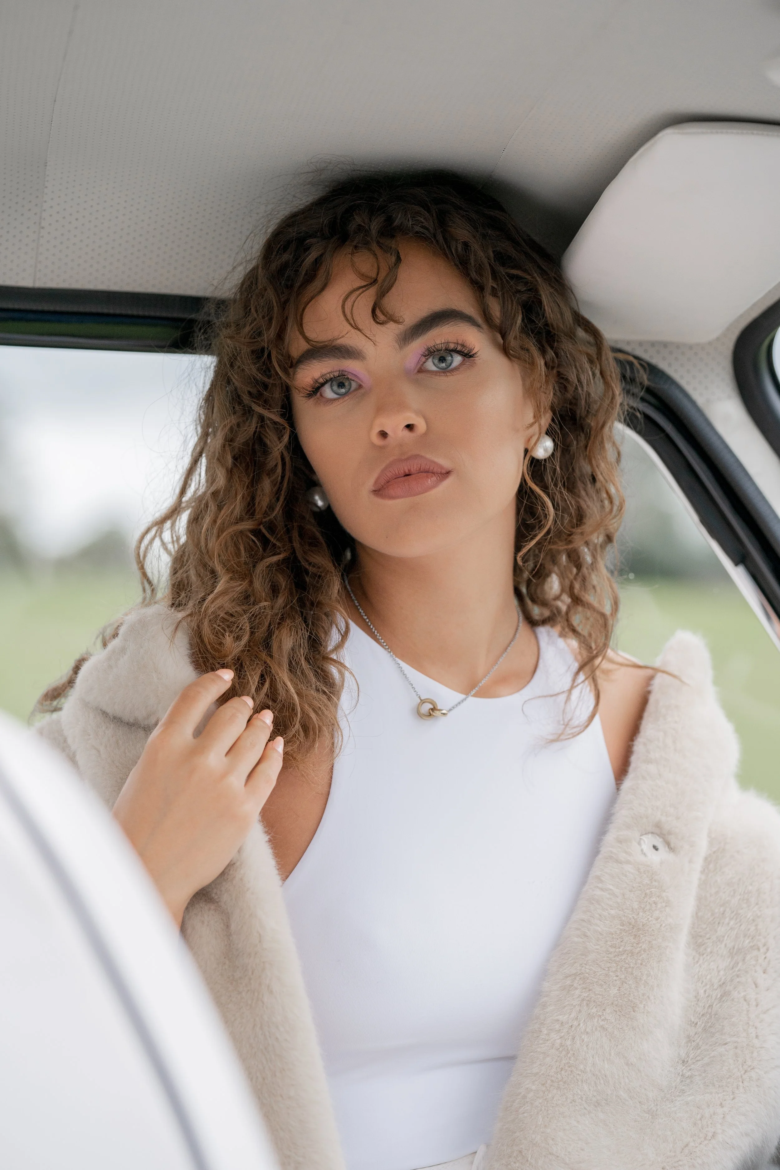 Une femme aux cheveux bouclés, portant un top blanc, une veste en peluche beige, des perles aux oreilles, et un collier en forme de boucle, regarde directement l'appareil photo dans l'intérieur d'une voiture.