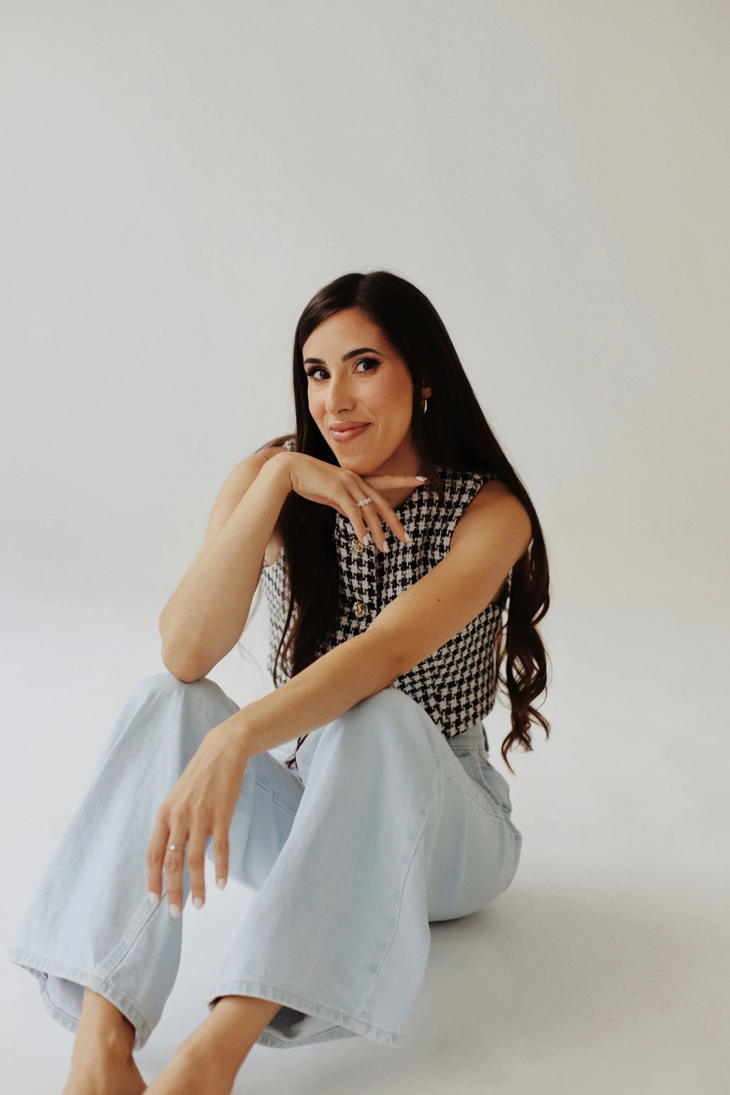 Femme assise sur le sol, souriante, portant un haut à carreaux noir et blanc et des jeans lâches, avec un fond blanc