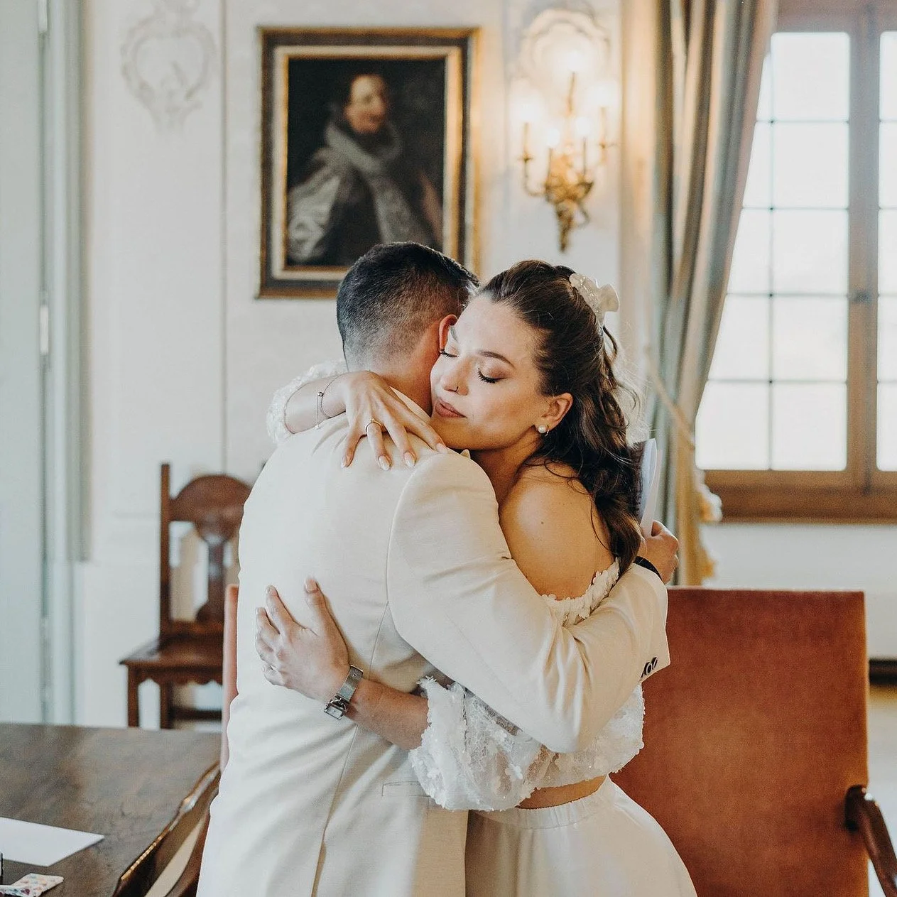 Deux personnes en habits de mariage s'étreignent dans une pièce élégante avec un portrait ancien en arrière-plan.