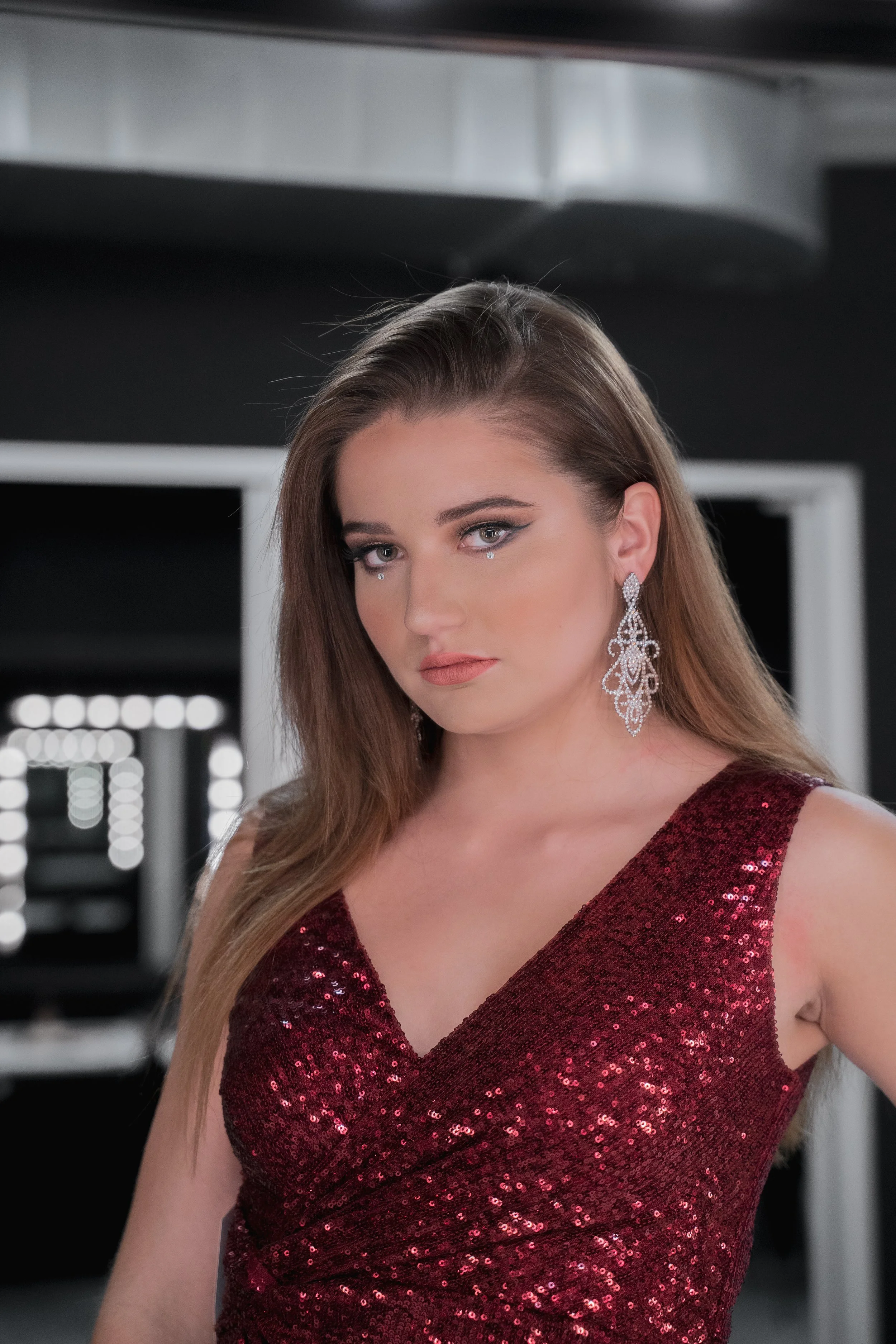 Jeune femme portant une robe rouge à sequins et de grands boucles d'oreilles, posant dans une pièce sombre avec miroir et lumières en arrière-plan.