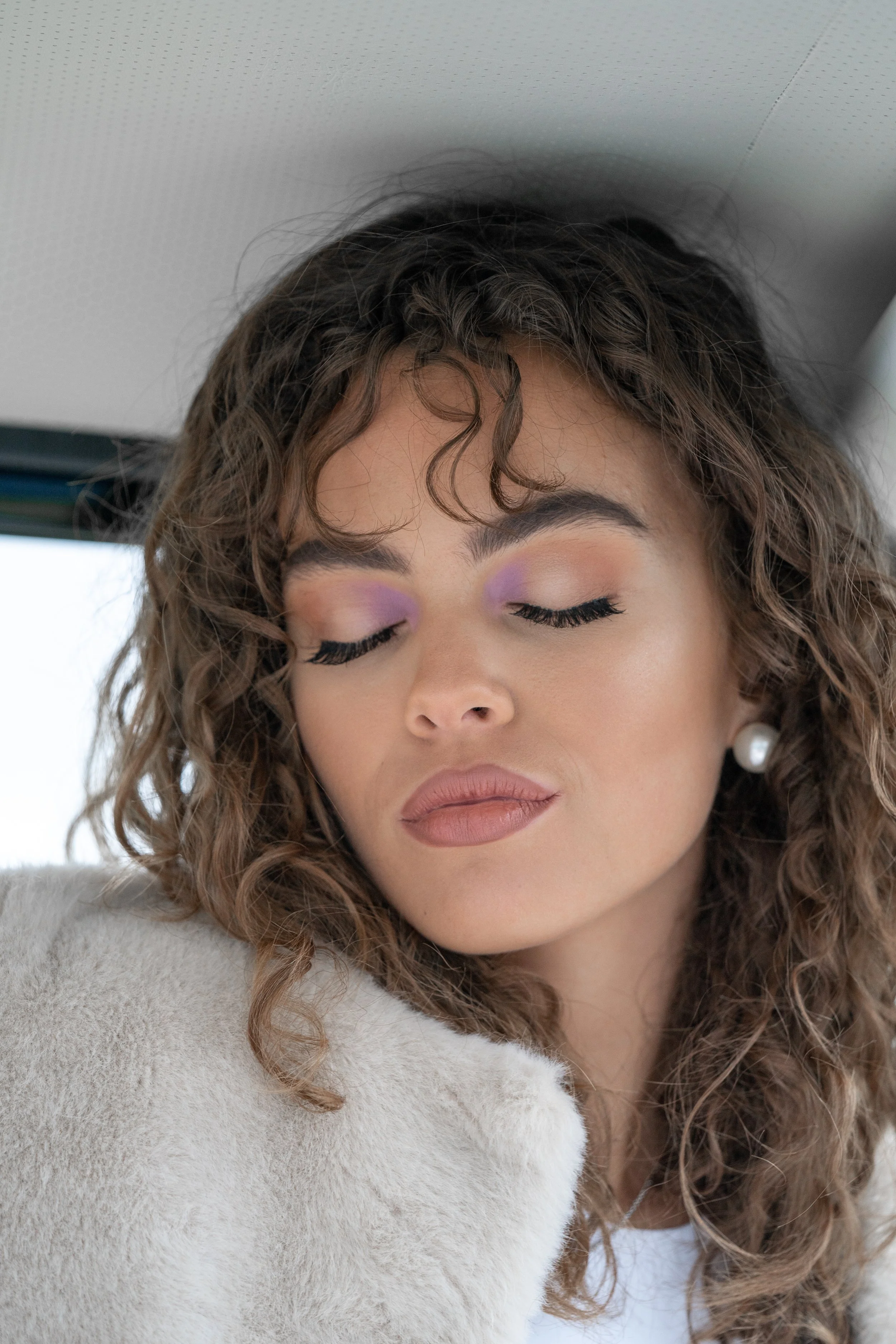 Femme avec maquillage violet clair, yeux fermés, portant une veste blanche en peluche et une boucle d'oreille perle, dans un intérieur moderne.
