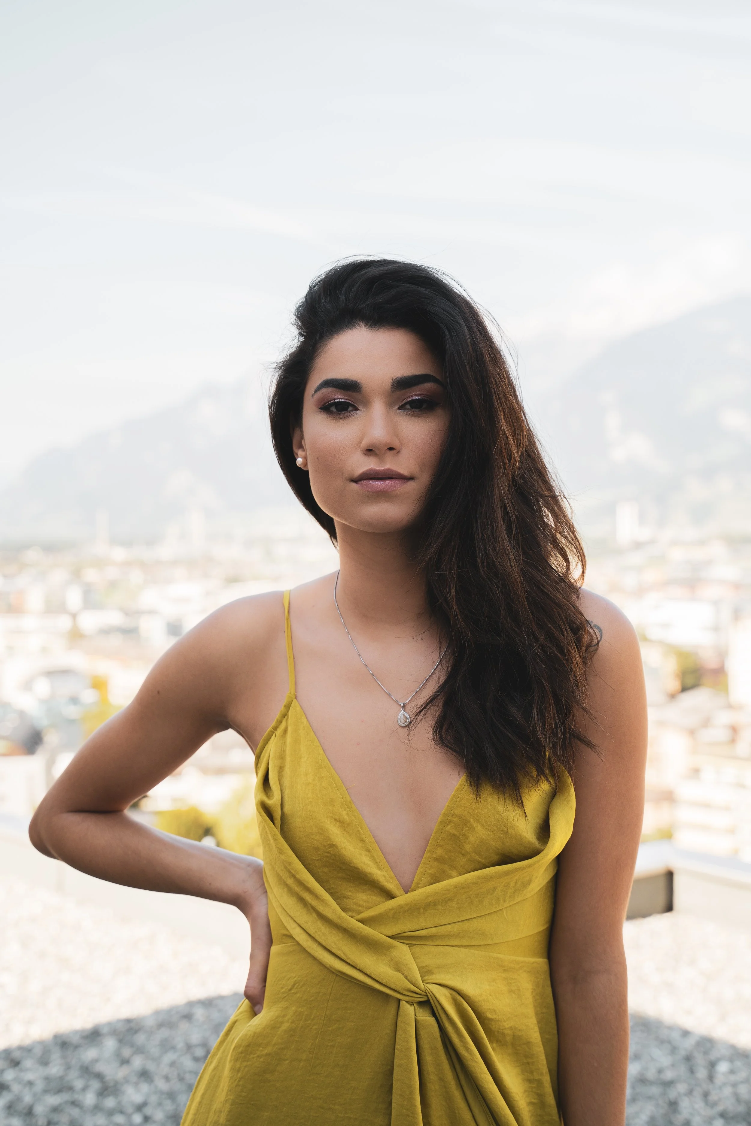 Jeune femme avec une robe jaune, cheveux longs et bruns, portant des boucles d'oreilles et un collier, se tenant devant une ville avec montagnes en arrière-plan.