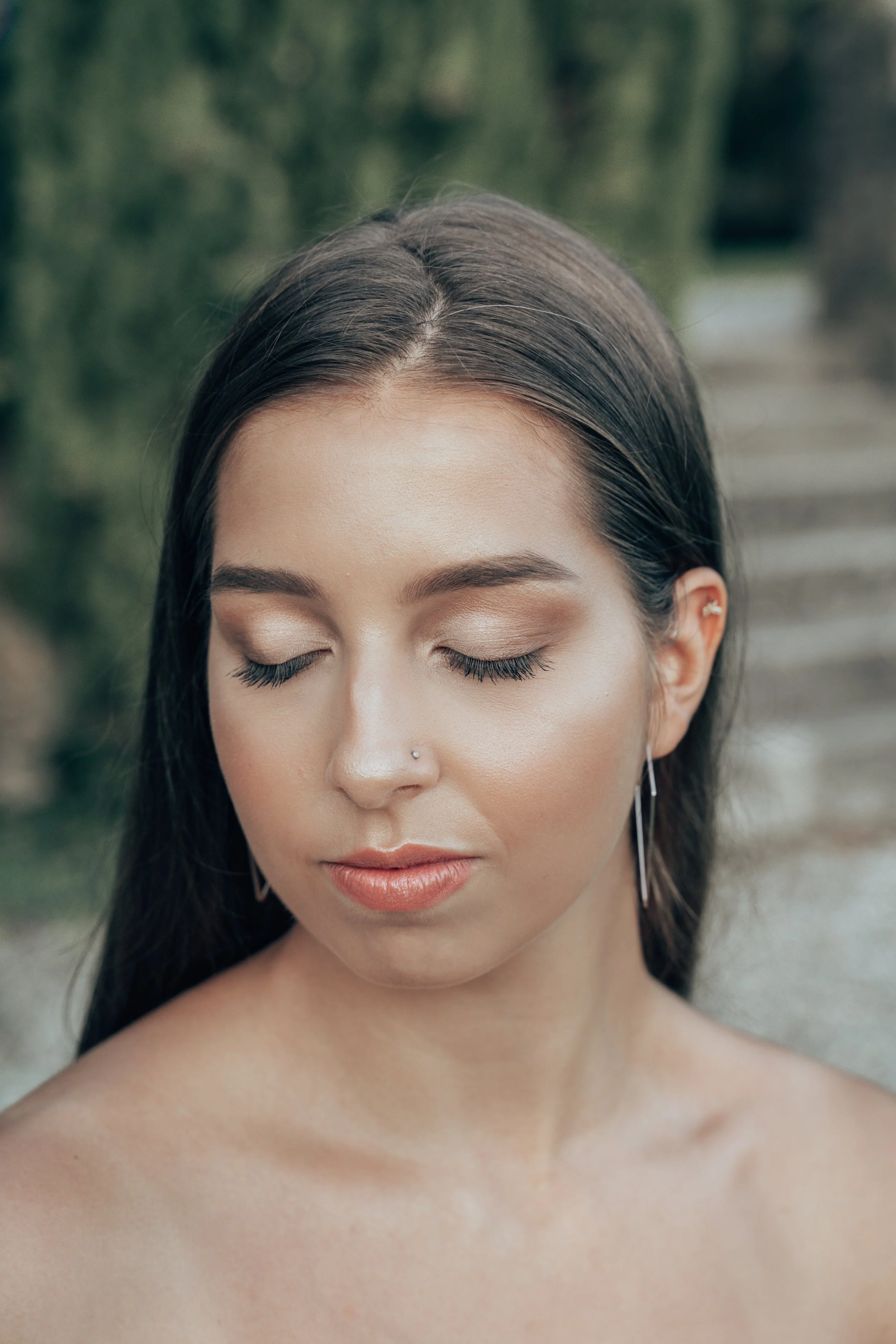 Jeune femme avec maquillage naturel, portant boucle d'oreille pendante, à l'extérieur, cheveux noirs longs, avec un fond flou de verdure et de sentiers.