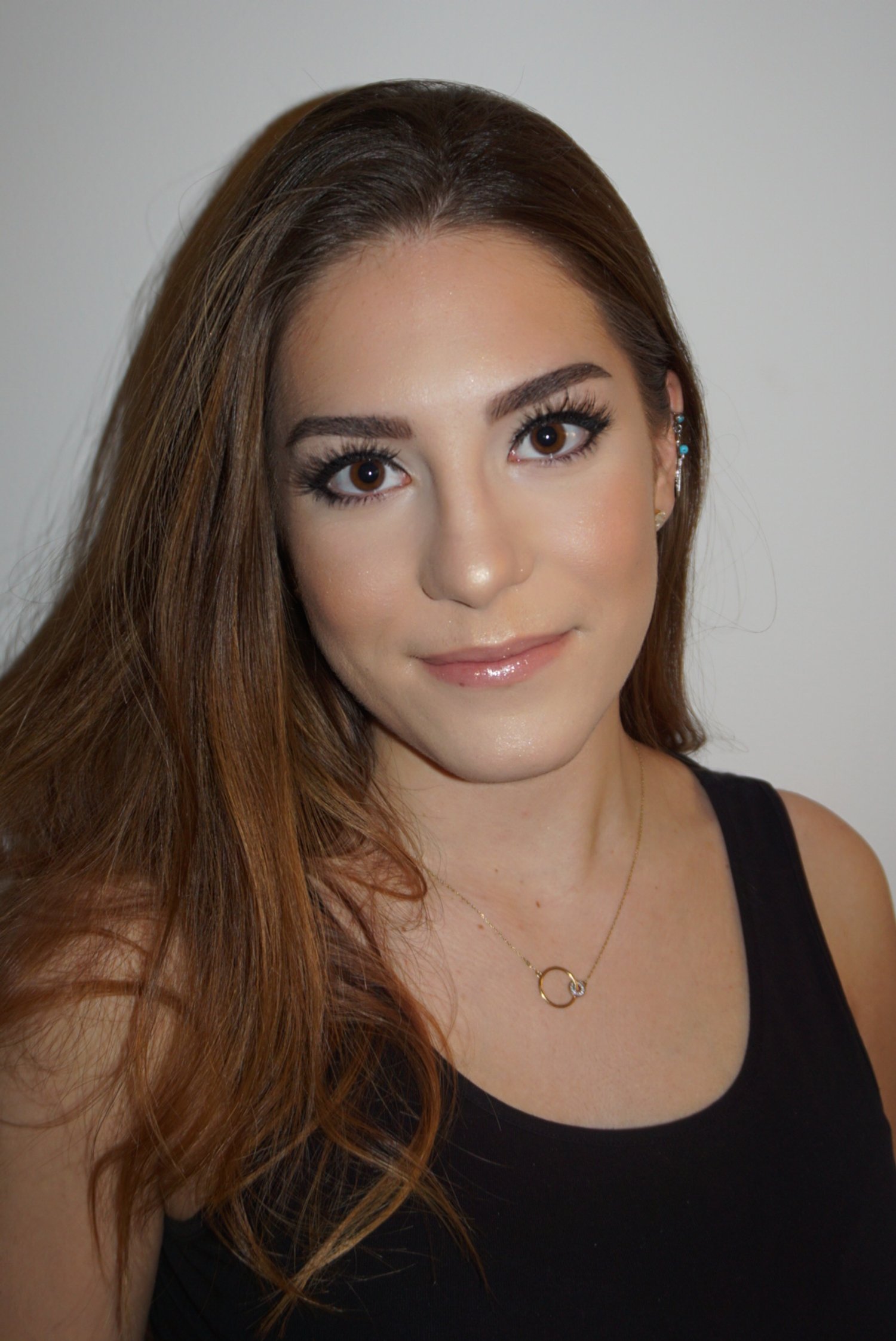 Portrait d'une femme aux cheveux bruns longs, souriante, portant un haut noir, un collier doré et des boucles d'oreilles bleues, devant un fond blanc.