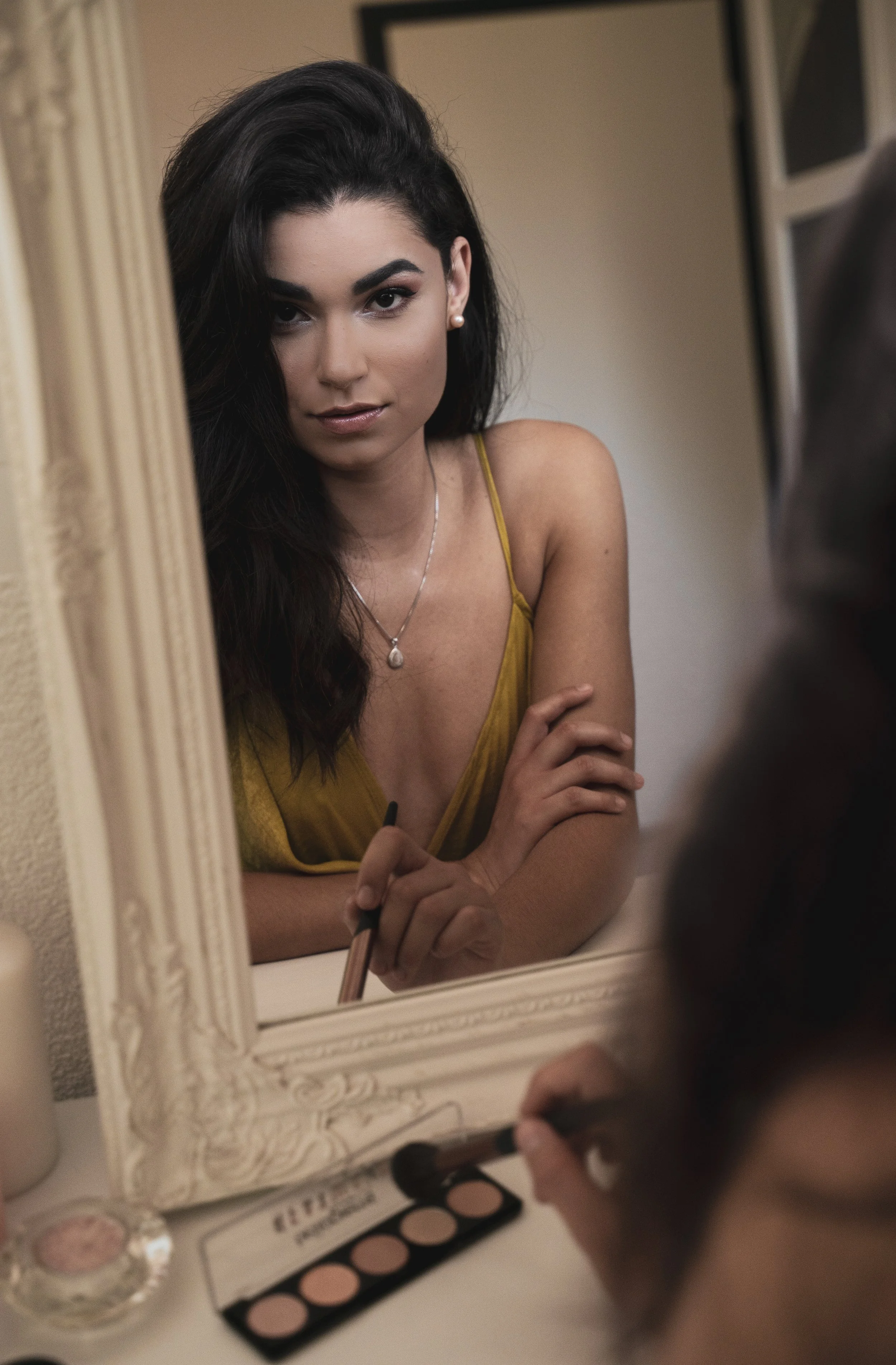 Une femme consulte un miroir en se maquillant, tenant un pinceau, avec une palette de maquillage sur la table devant elle.