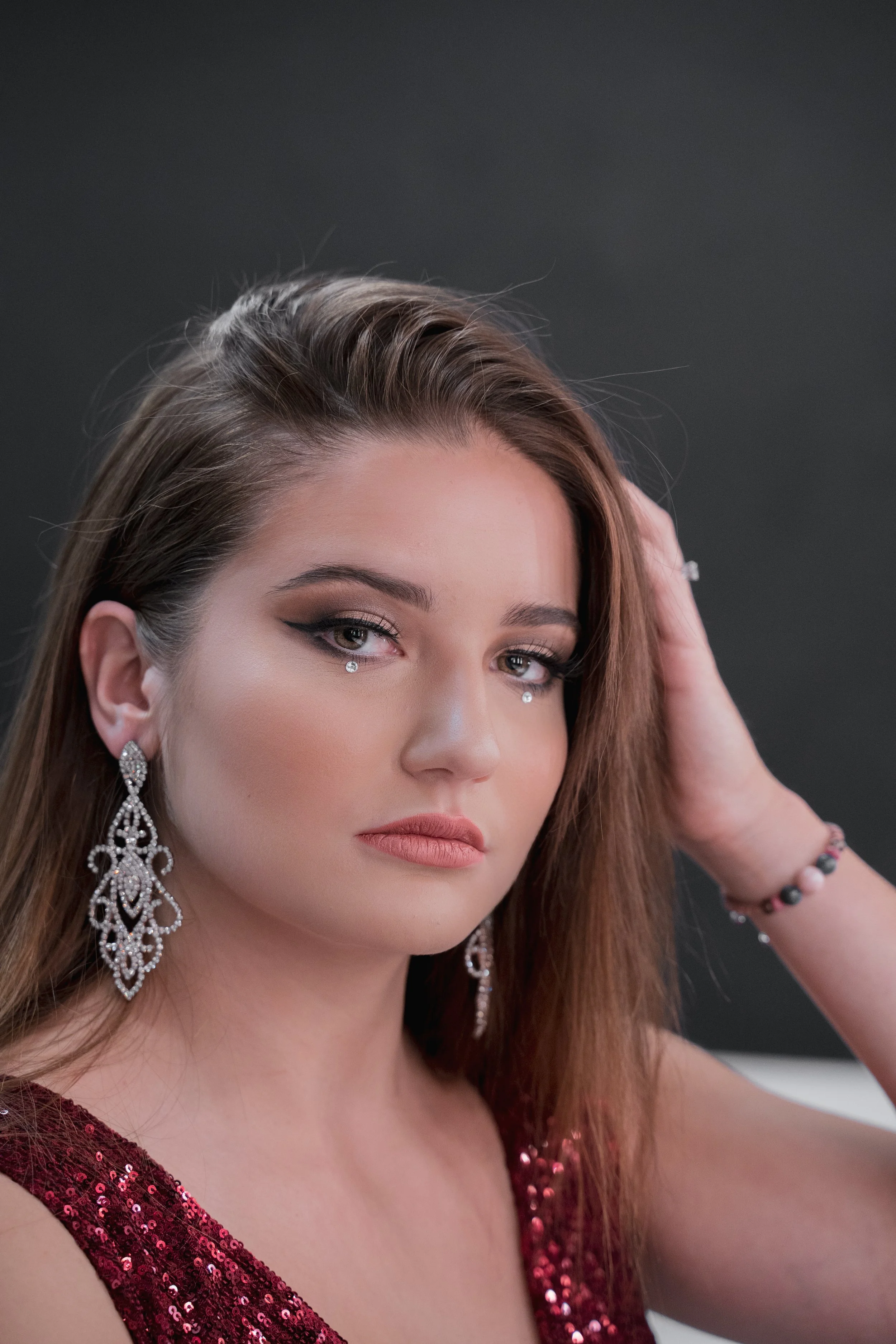 Portrait d'une femme avec maquillage sophistiqué, portant de grandes boucles d'oreilles en diamant, un bracelet en perles, et une robe ornée de sequins rouges, tenant sa tête d'une main contre un fond sombre.
