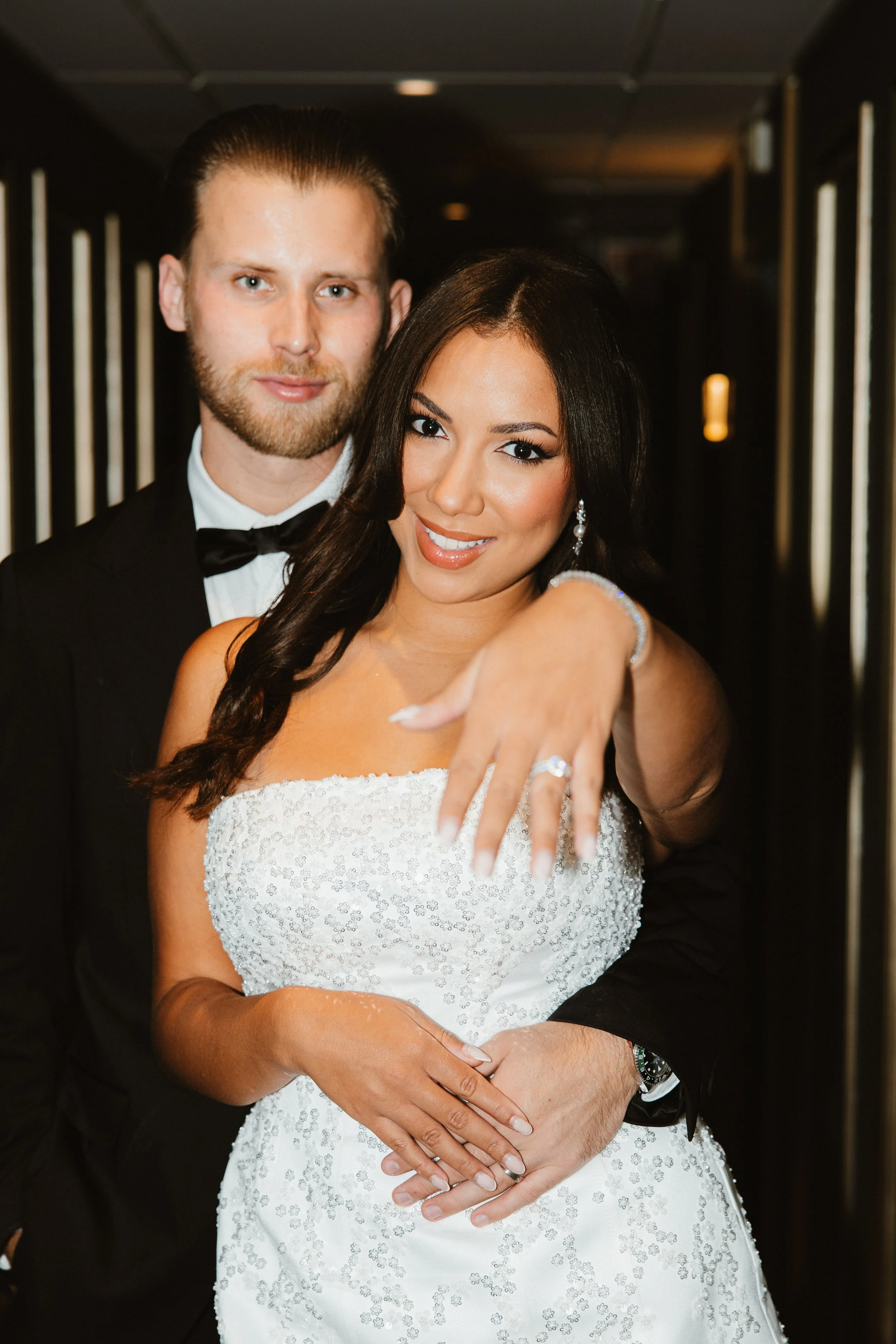 Un couple en robe de mariée et costume de mariage posant dans un couloir, la femme montrant la bague de mariage à la caméra.