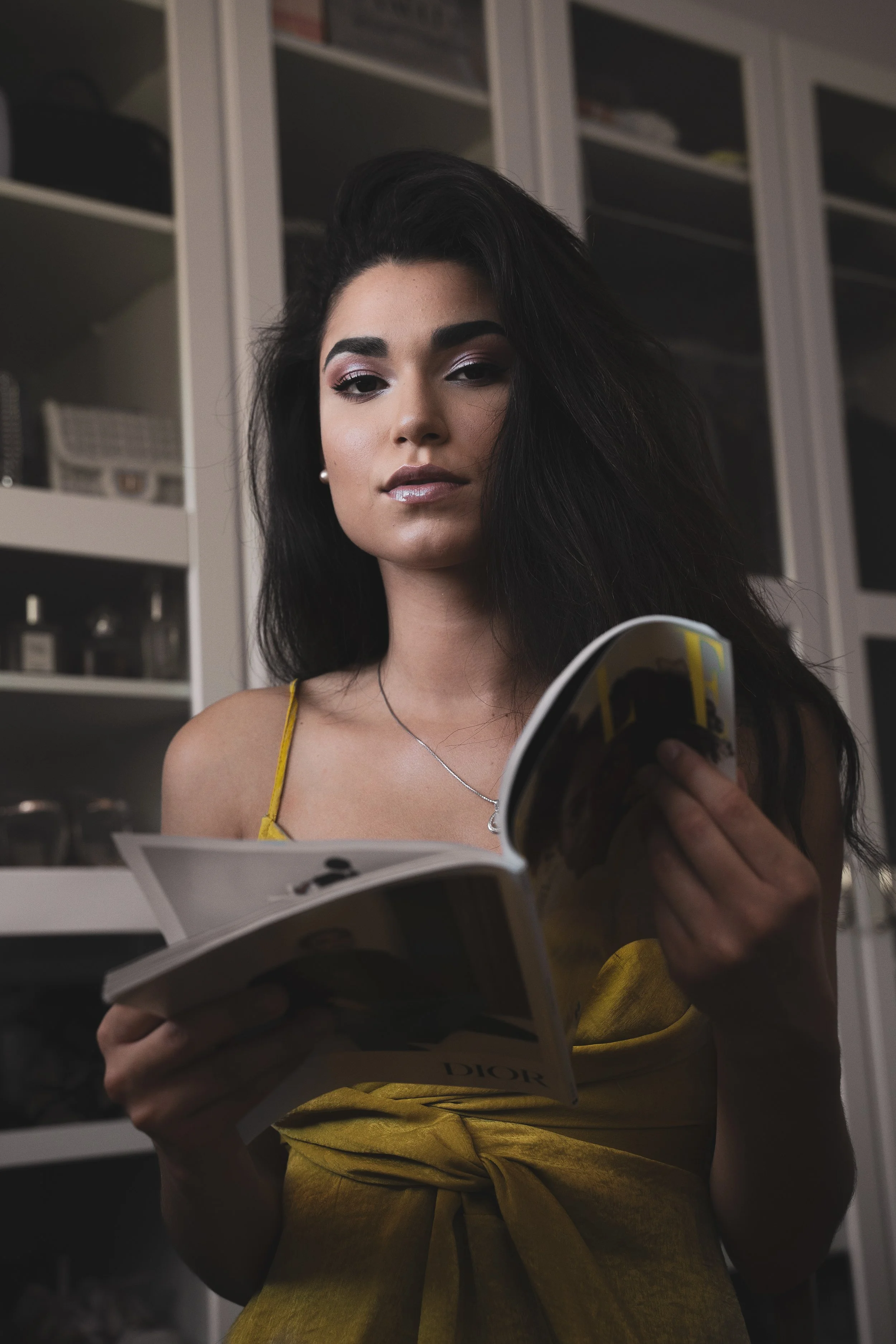 Une femme aux cheveux noirs longs, portant un haut jaune à baillettes, regarde un magazine dans un intérieur moderne avec des armoires en arrière-plan.