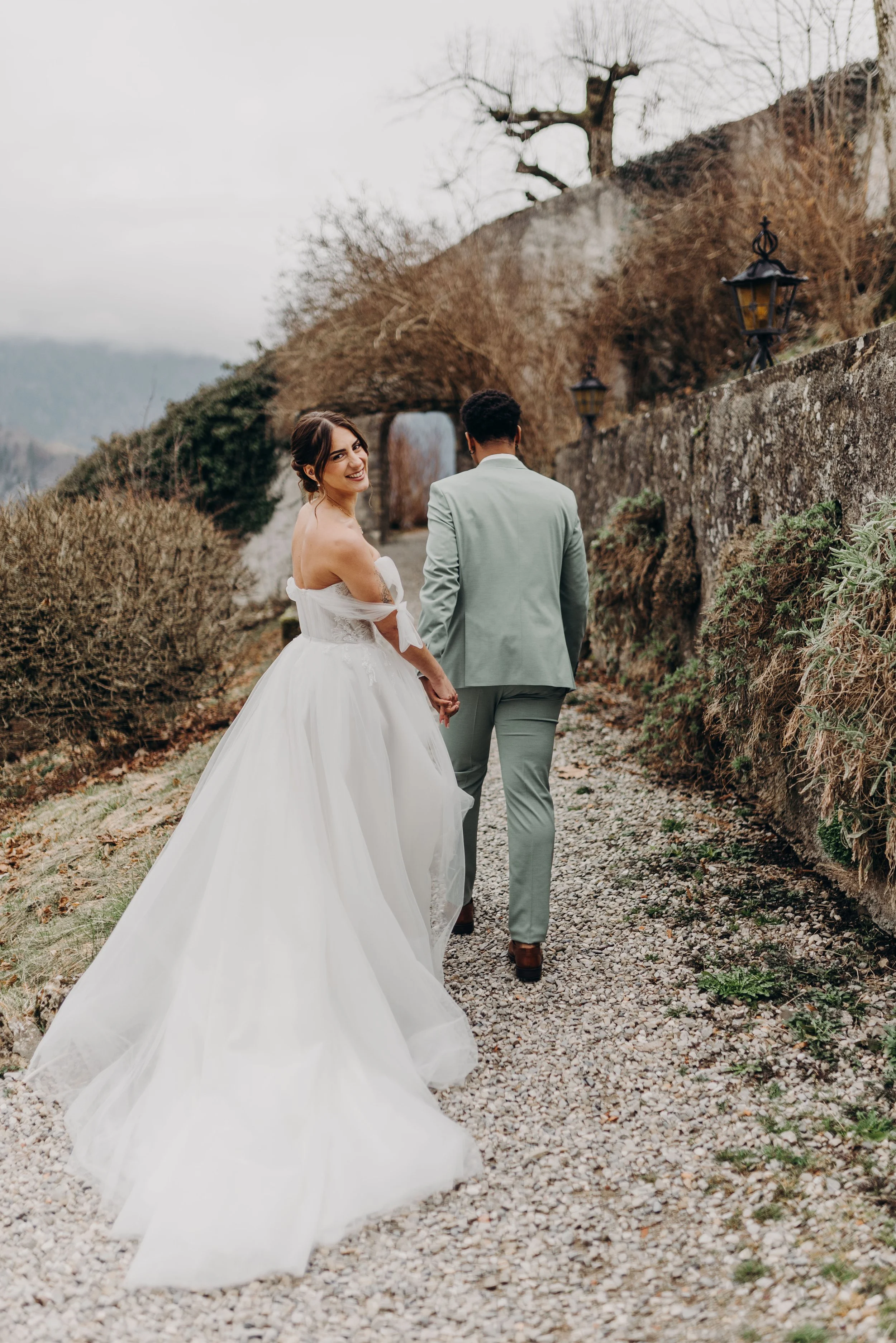 Un couple de mariage marche main dans la main sur un chemin en plein air, la femme sourit et porte une robe de mariée blanche, l'homme porte un costume gris clair, l'arrière-plan montre un paysage naturel avec un mur en pierre, des lampadaires et un arbre dénudé.