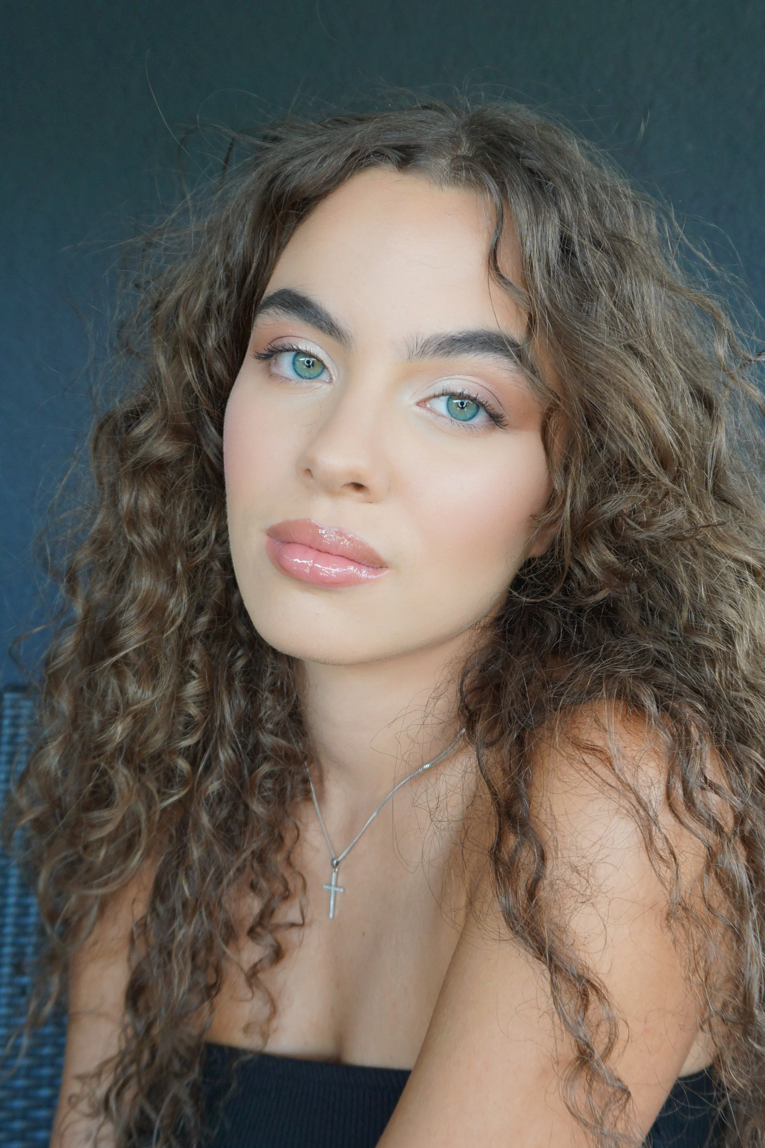 Un portrait d'une jeune femme aux yeux bleus, cheveux bouclés, portant un collier avec une croix, souriant légèrement contre un fond sombre.