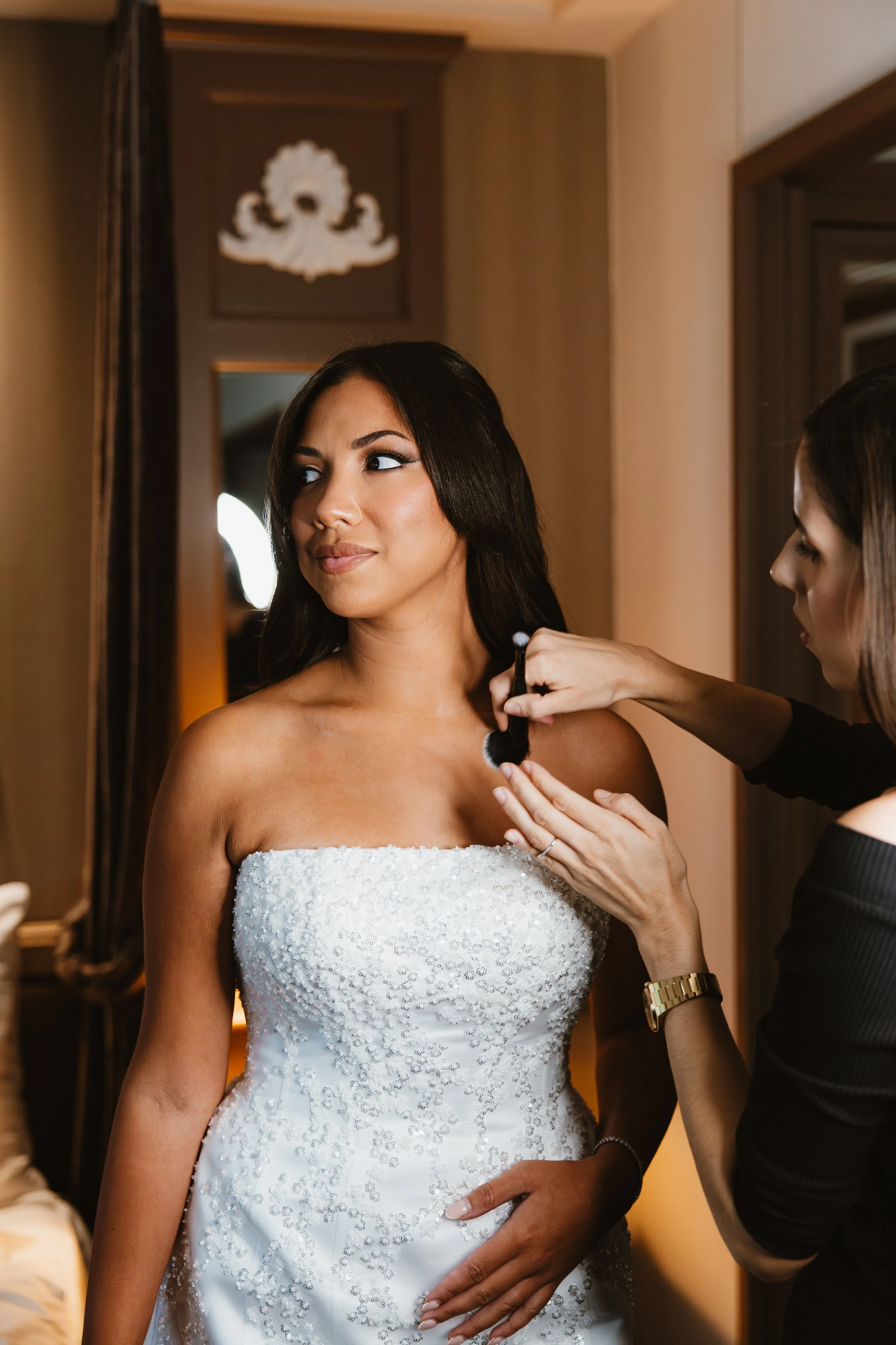Une femme portant une robe de mariée reçoit une retouche de maquillage ou de coiffure par une autre femme dans une pièce bien éclairée.