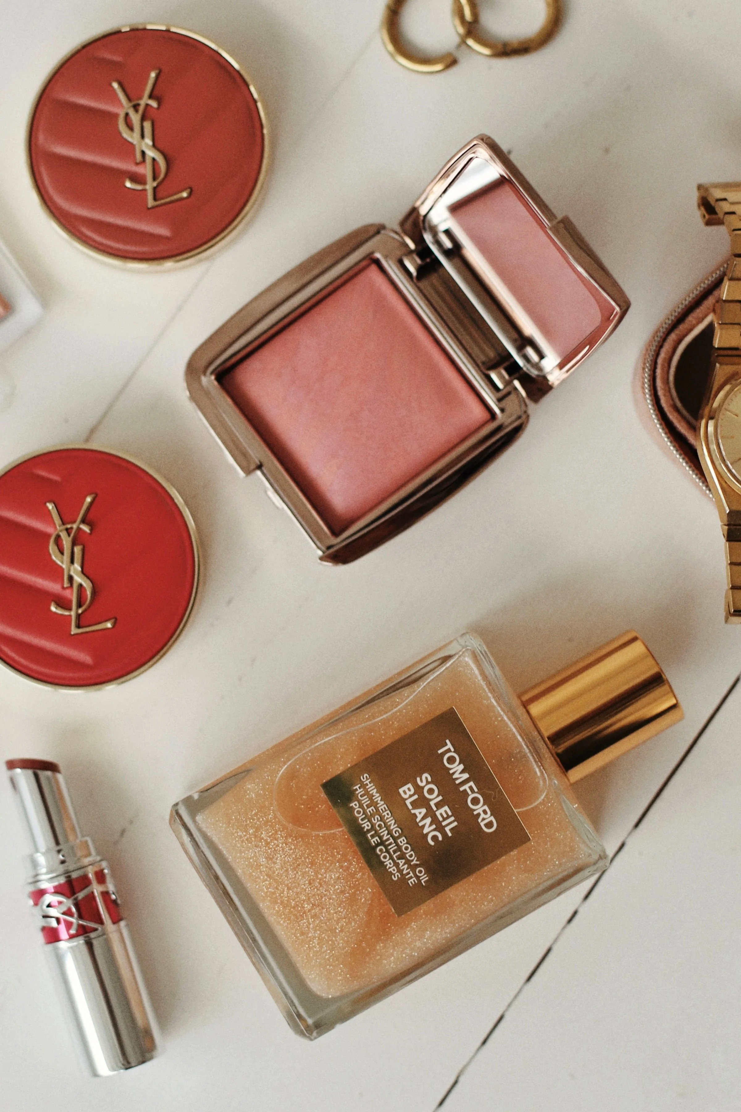 Collection de produits de beauté et accessoires, notamment deux poudres compactes Yves Saint Laurent, une bouteille de parfum Tom Ford Soleil Blanc, un rouge à lèvres Yves Saint Laurent, et une montre or.
