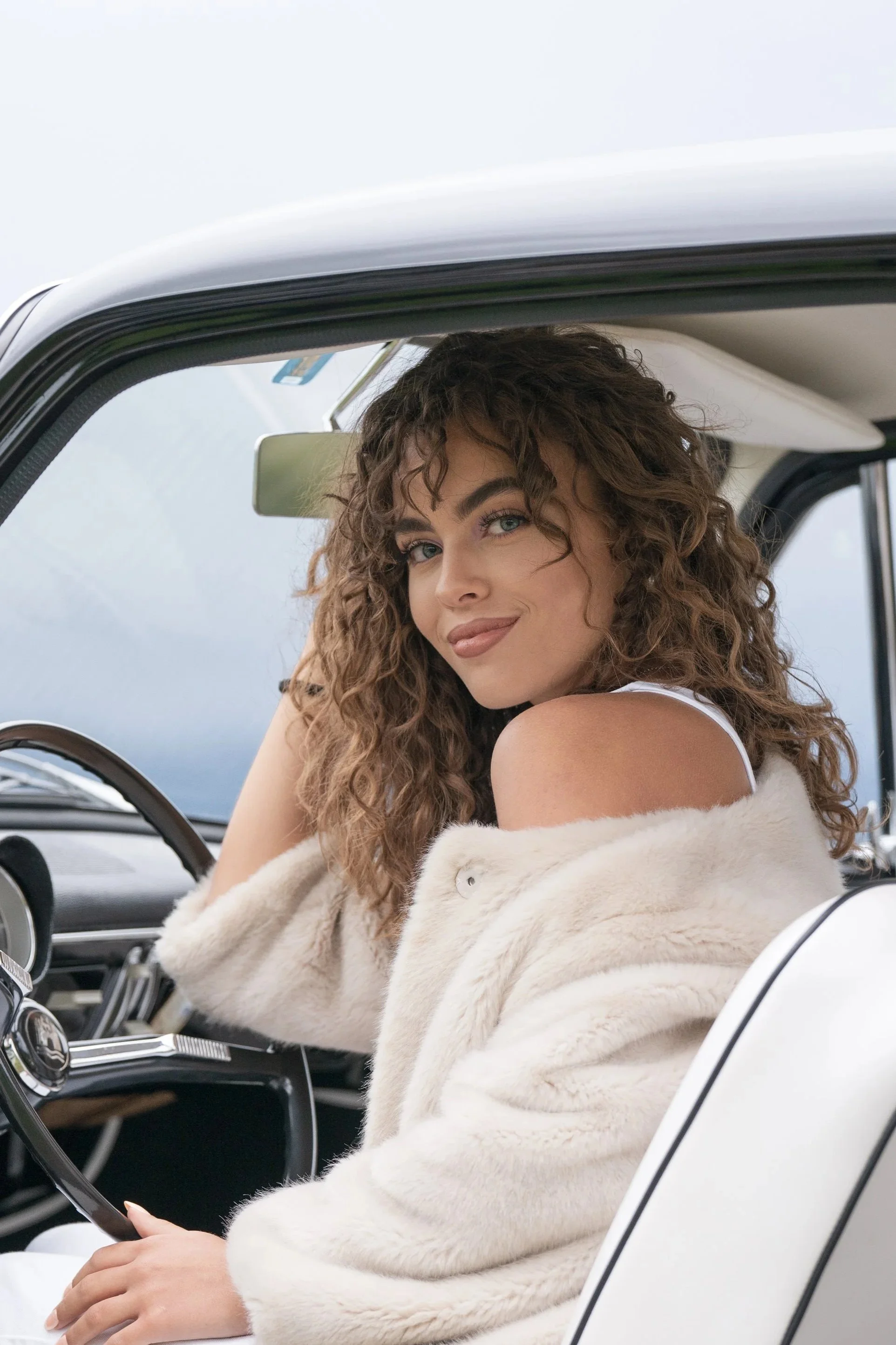 Femme aux cheveux marron bouclés assise dans une voiture, portant une veste en fourrure beige, regardant par la fenêtre avec un sourire.