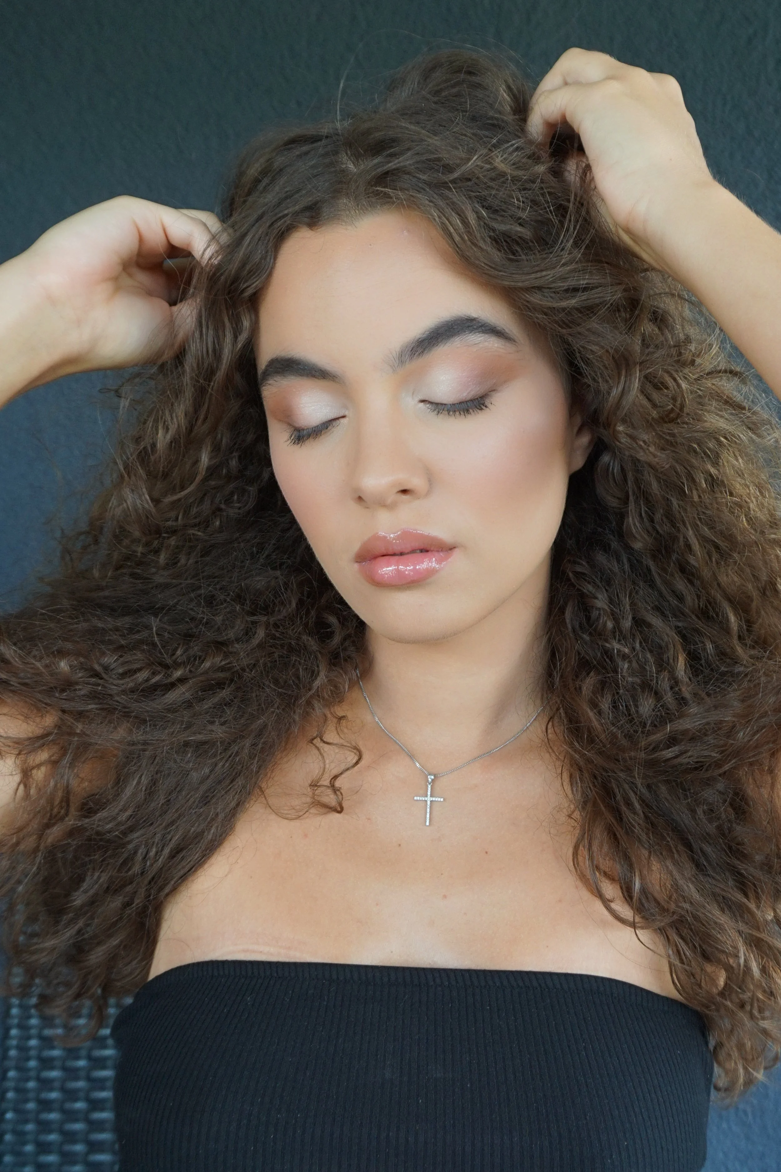 Jeune femme aux cheveux bouclés, yeux fermés, portant un collier avec une croix, vêtue d'un haut noir.Elle pose contre un fond gris.