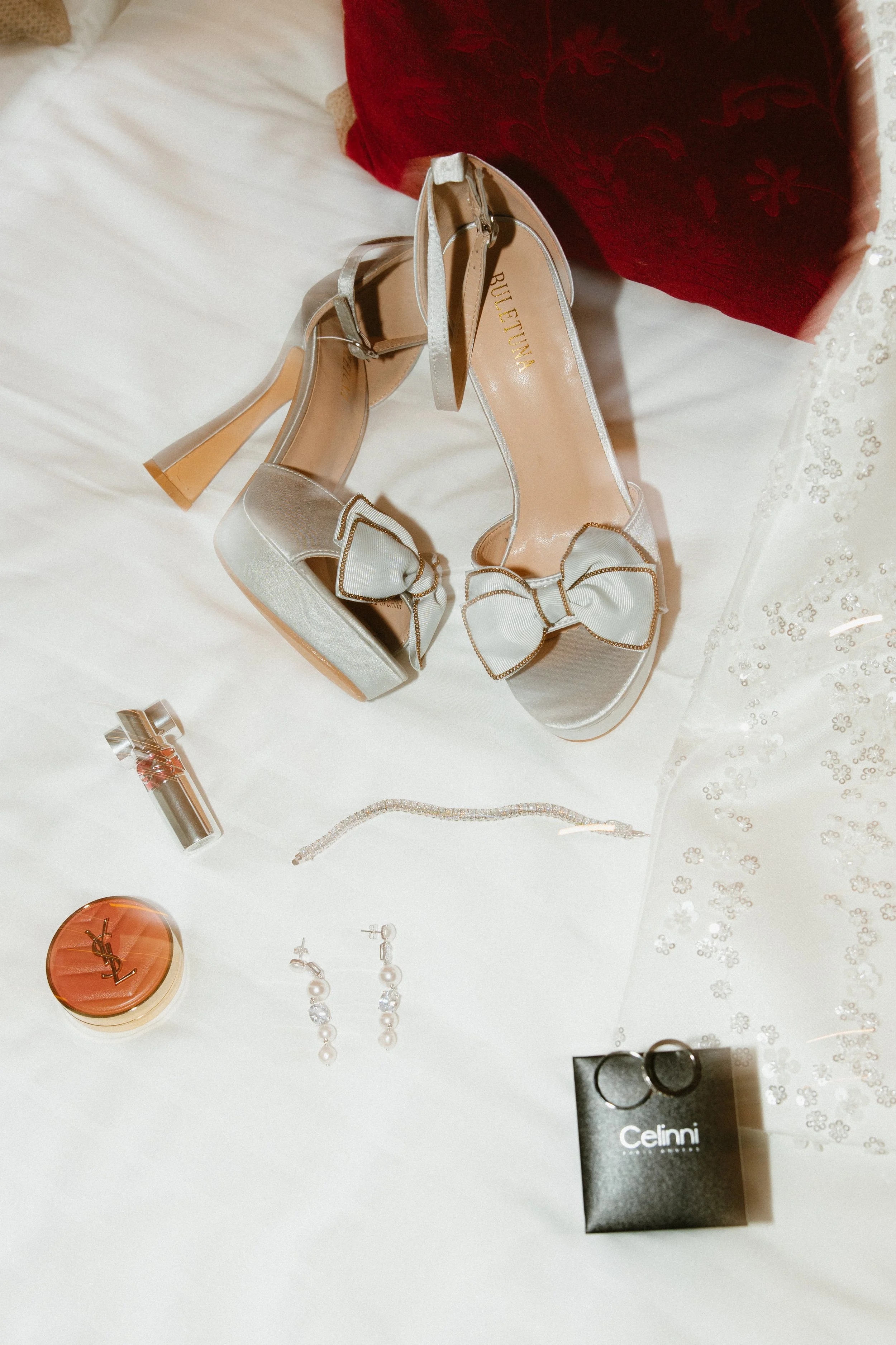 Chaussures à talons ornées de rubans, bijoux, parfum Yves Saint Laurent, bagues, et autres accessoires sur une toile blanche près d'un tissu en dentelle et d'un coussin rouge.