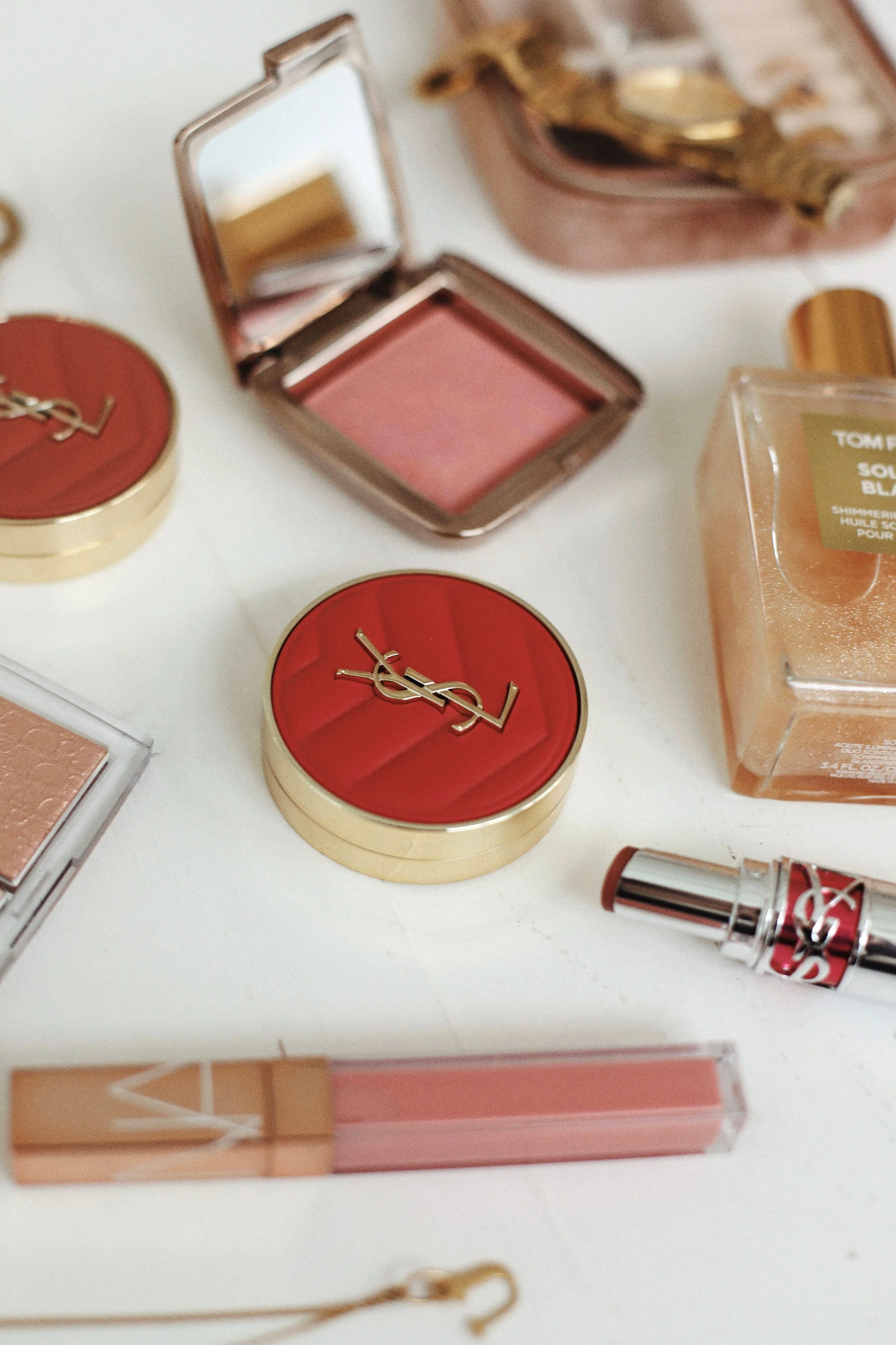 Collection de produits de maquillage, notamment un poudrier YSL rouge, un rouge à lèvres, des poudres et une bouteille de parfum.