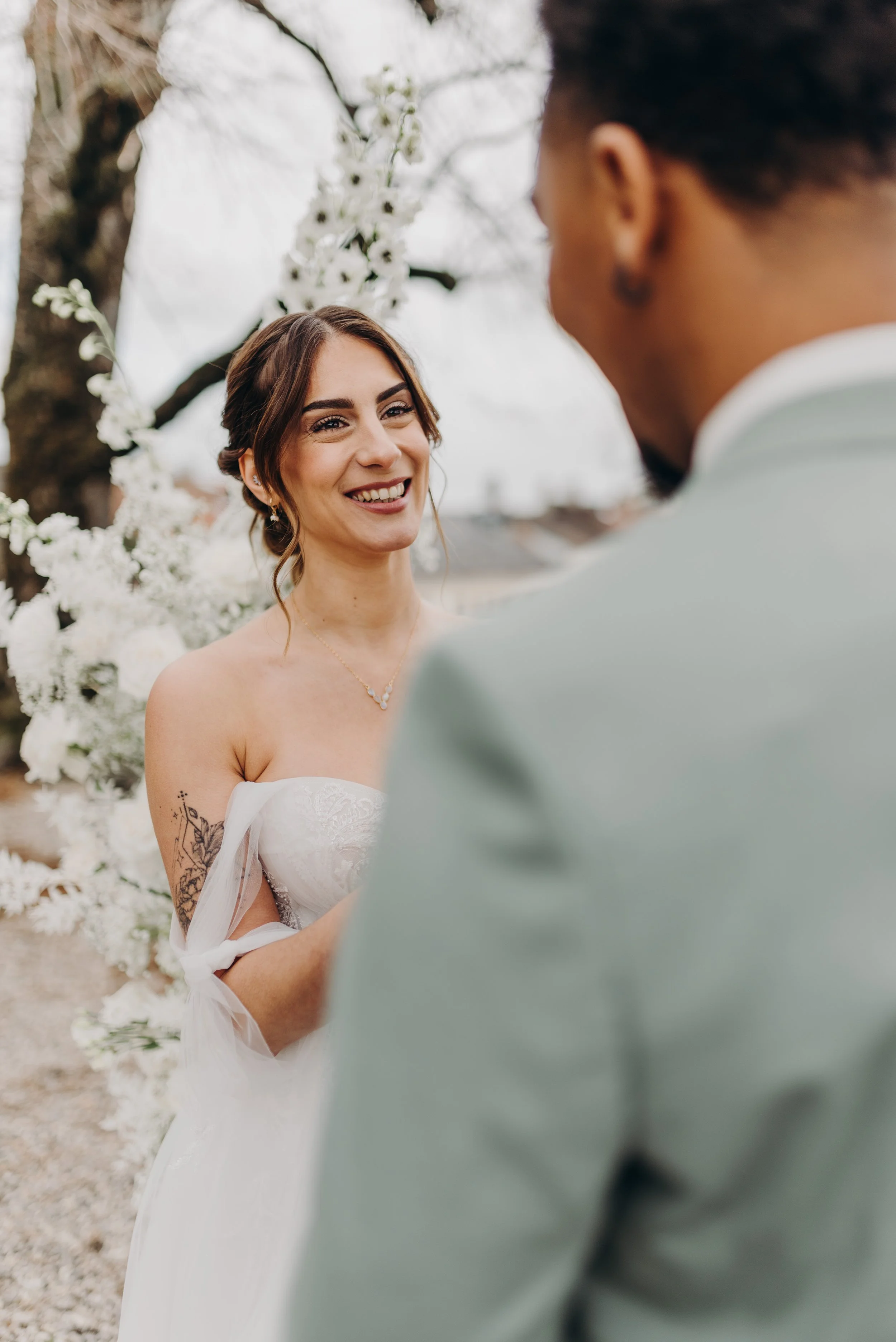 Une femme en robe de mariée sourit lors d'un mariage en extérieur. Elle porte un collier et a une tatouage sur le haut de son bras. En arrière-plan, il y a un décor floral avec des fleurs blanches et un arbre.