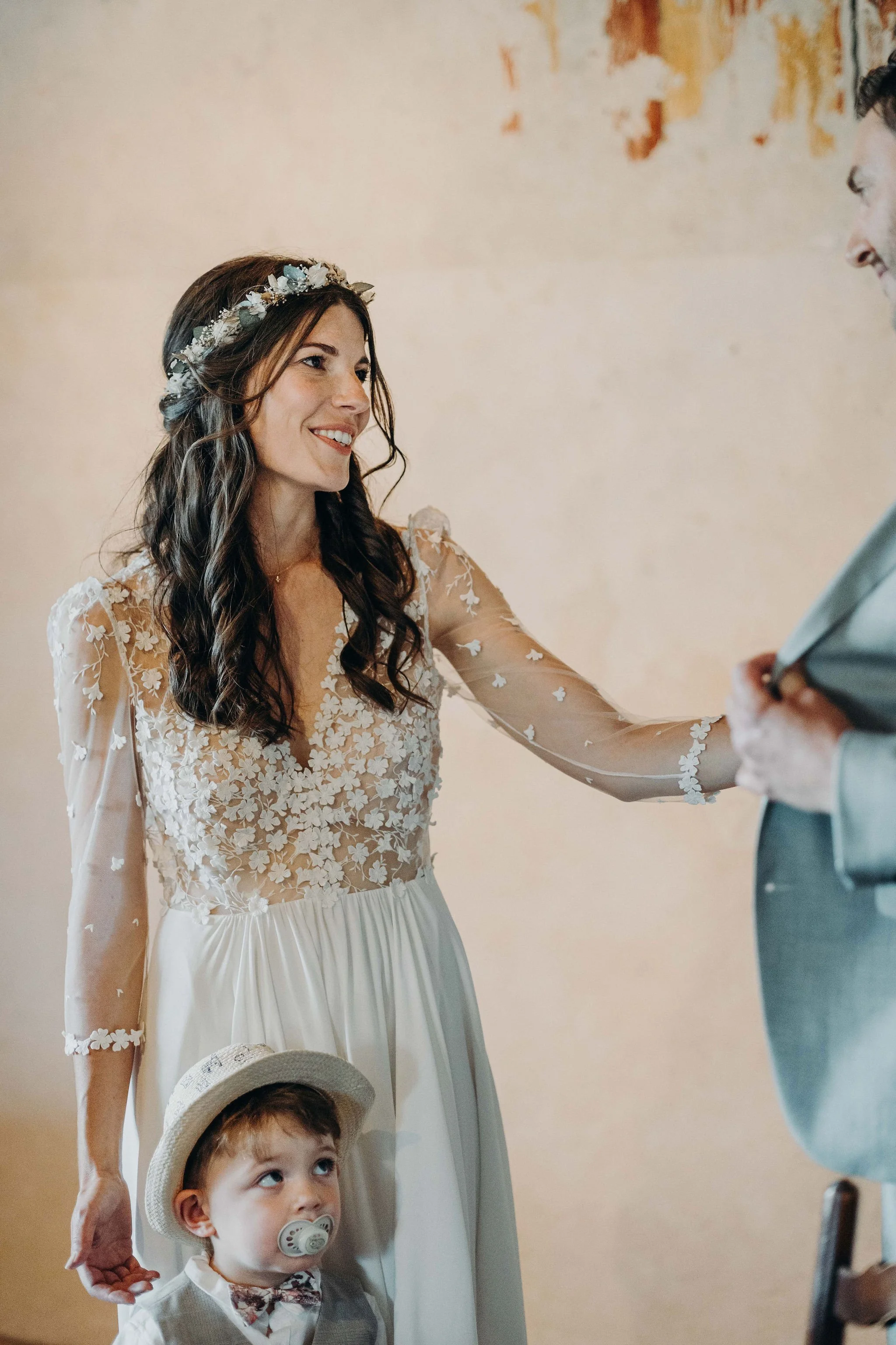 Une femme souriante en robe de mariage avec une couronne de fleurs, tenant un jeune garçon avec un tétine, lors d'une cérémonie de mariage.