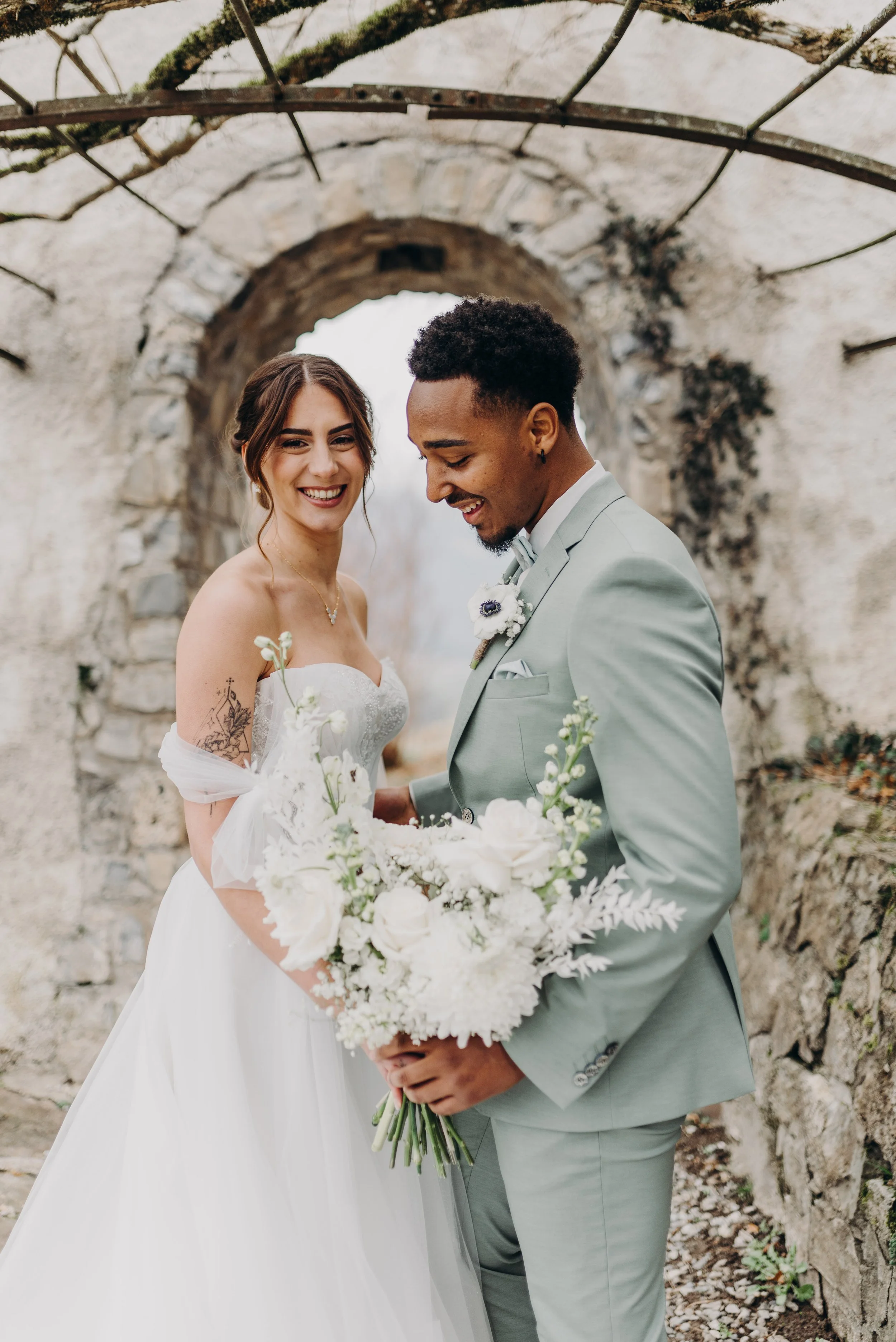 Un couple de mariés souriants, devant un vieux mur en pierre, tenant un bouquet de fleurs blanches lors d'un mariage.