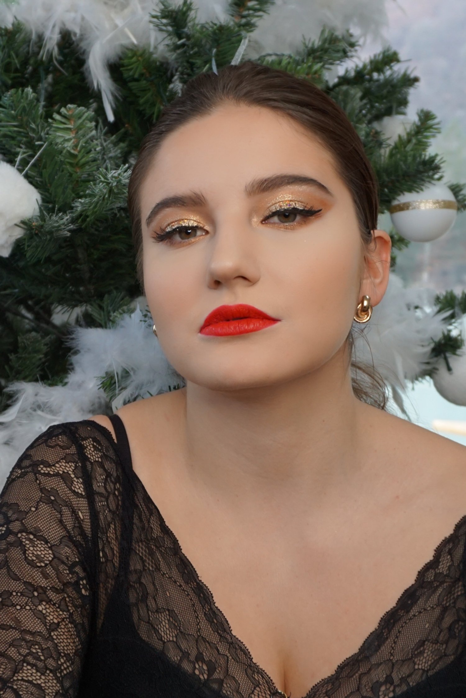 Une femme aux cheveux bruns, maquillage sophistiqué avec yeux dorés et lèvres rouges, portant un haut en dentelle noire, posant devant un sapin de Noël décoré.