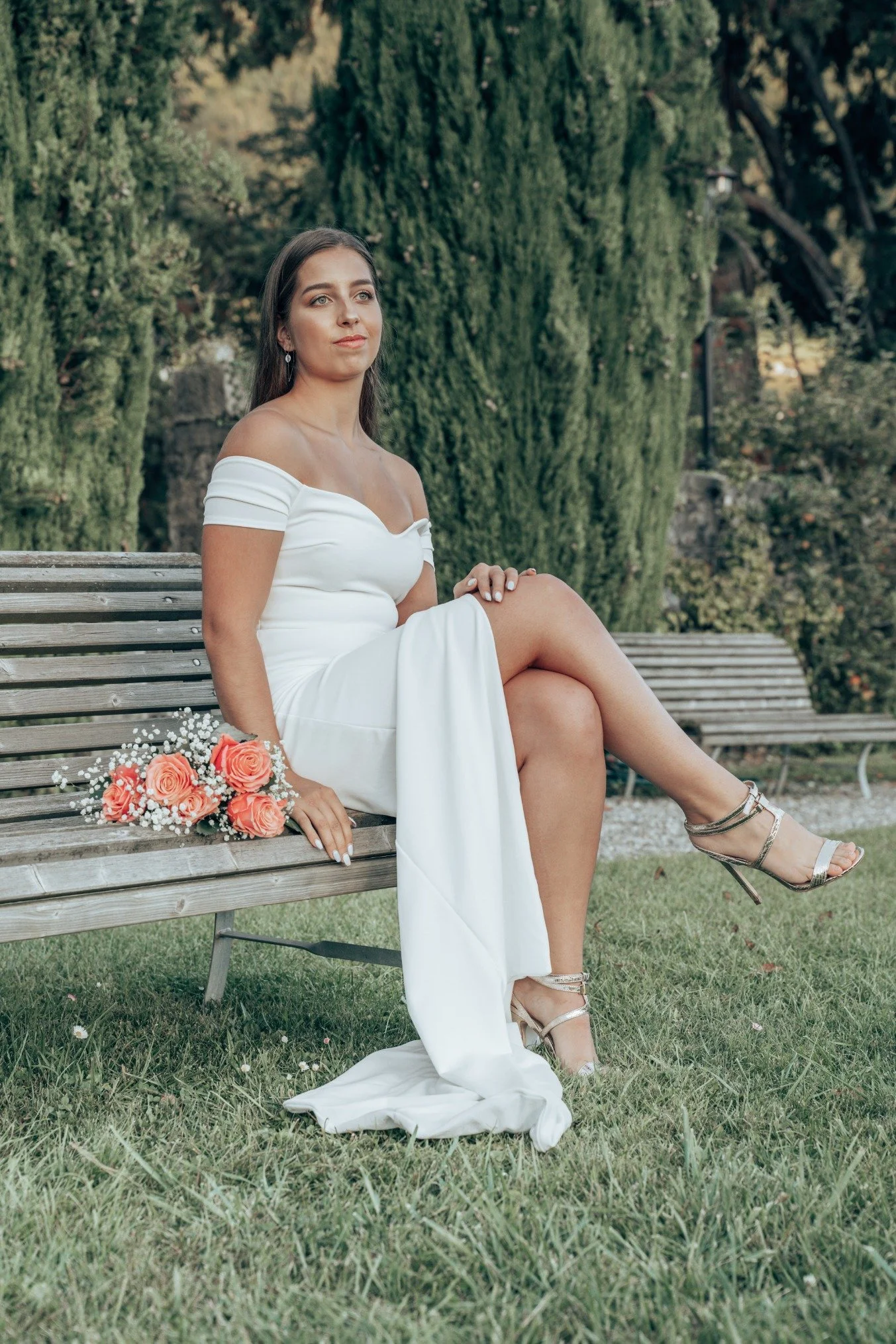 Femme en robe de mariage assise sur un banc en bois, avec un bouquet de roses, dans un parc avec des arbres verts.