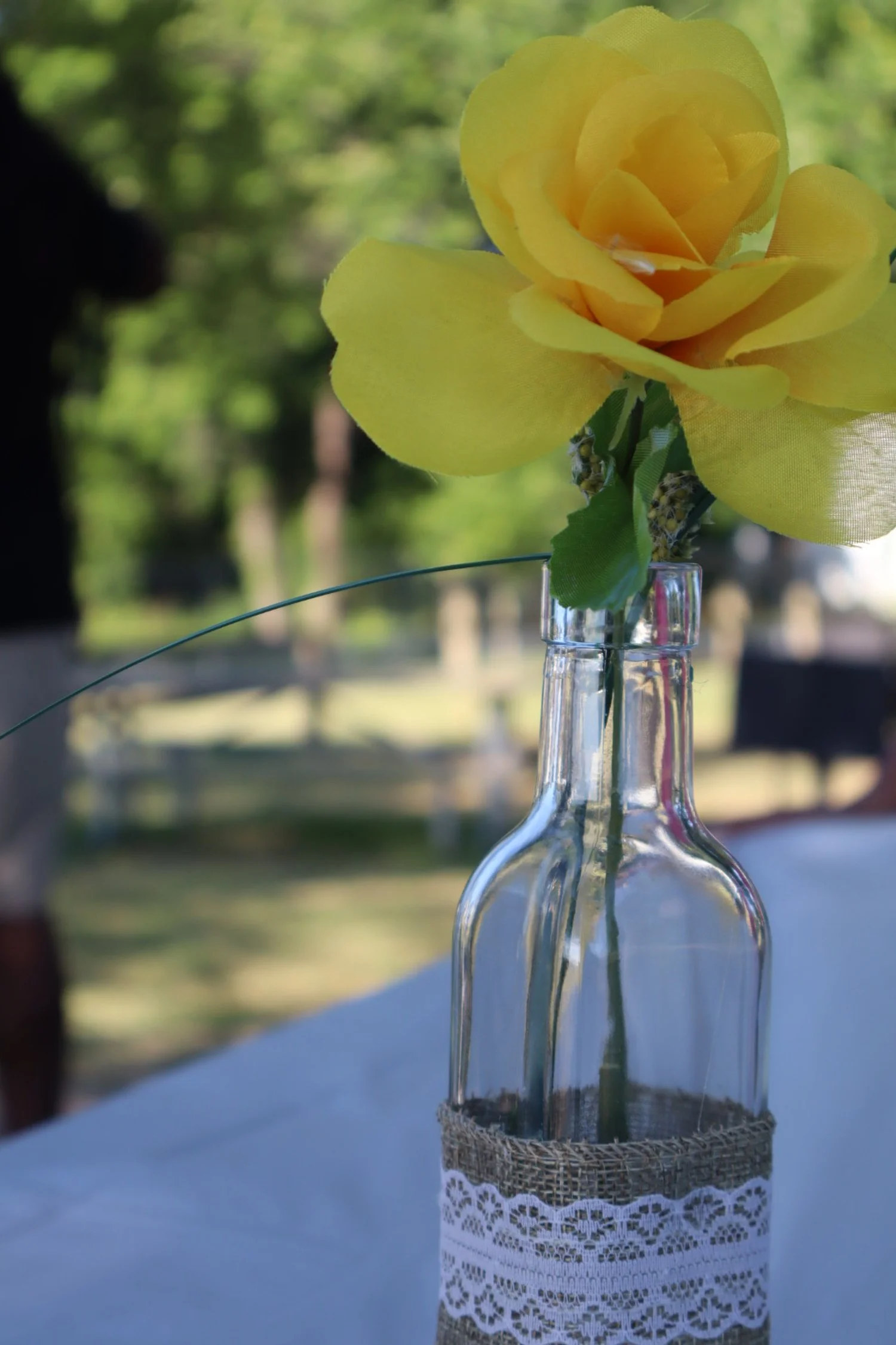 Yellow artificial rose in a glass bottle with lace and burlap fabric, outdoors with blurred green trees in the background.