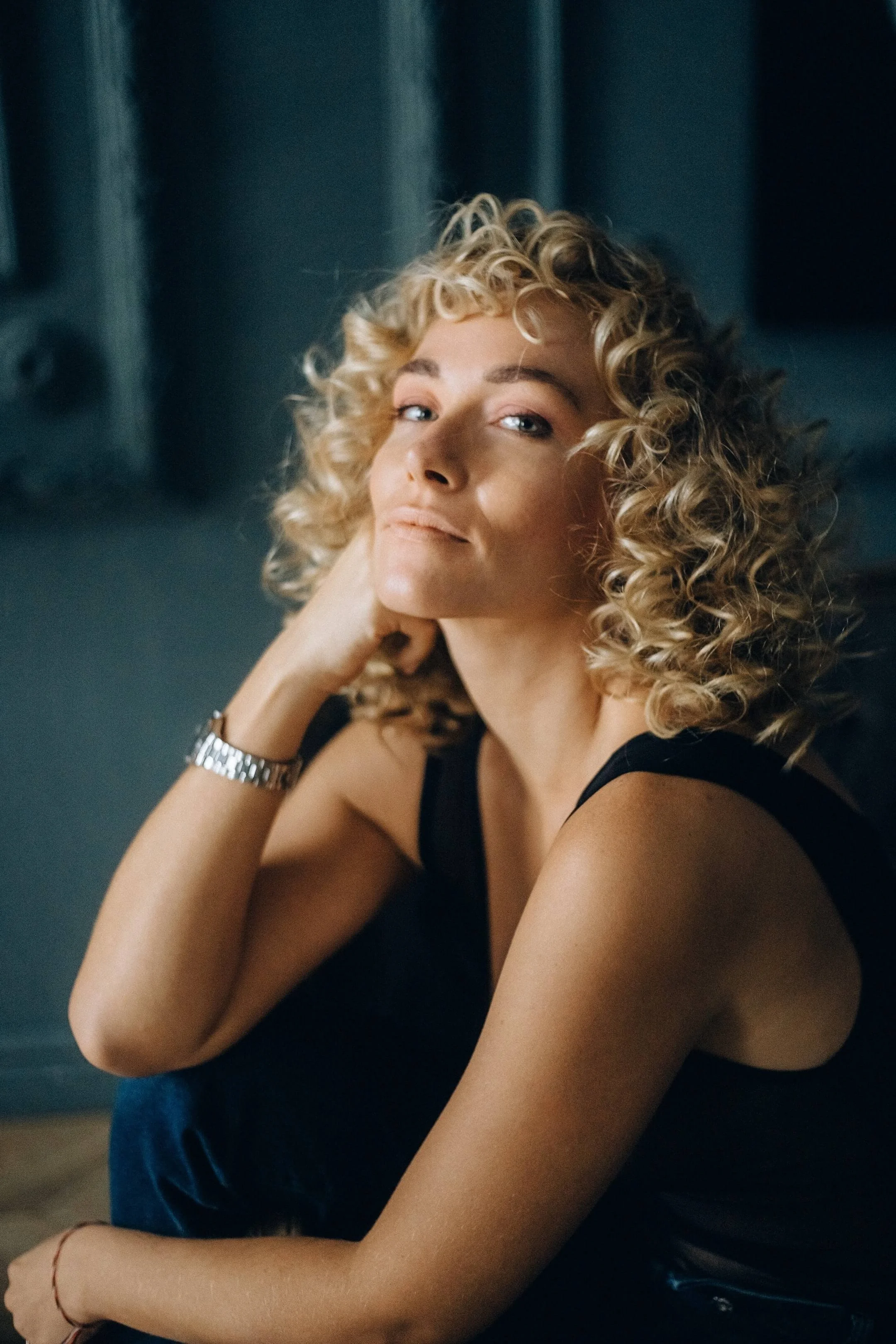 A woman with curly blonde hair gazes at the camera, resting her chin on her hand, wearing a black sleeveless top and a watch.