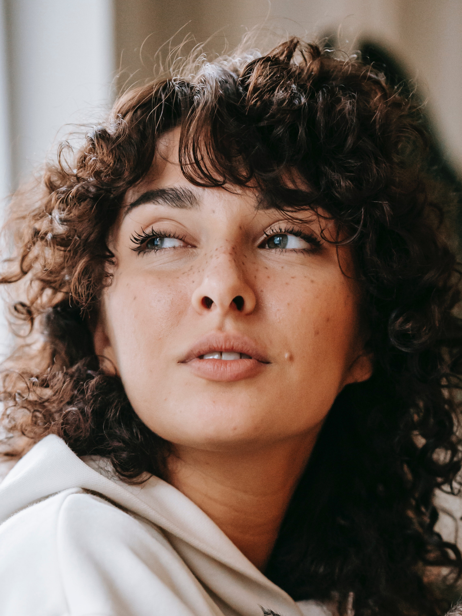 Close-up of a woman with curly brown hair, blue eyes, and freckles, looking to the side with a gentle expression, wearing a white hoodie.