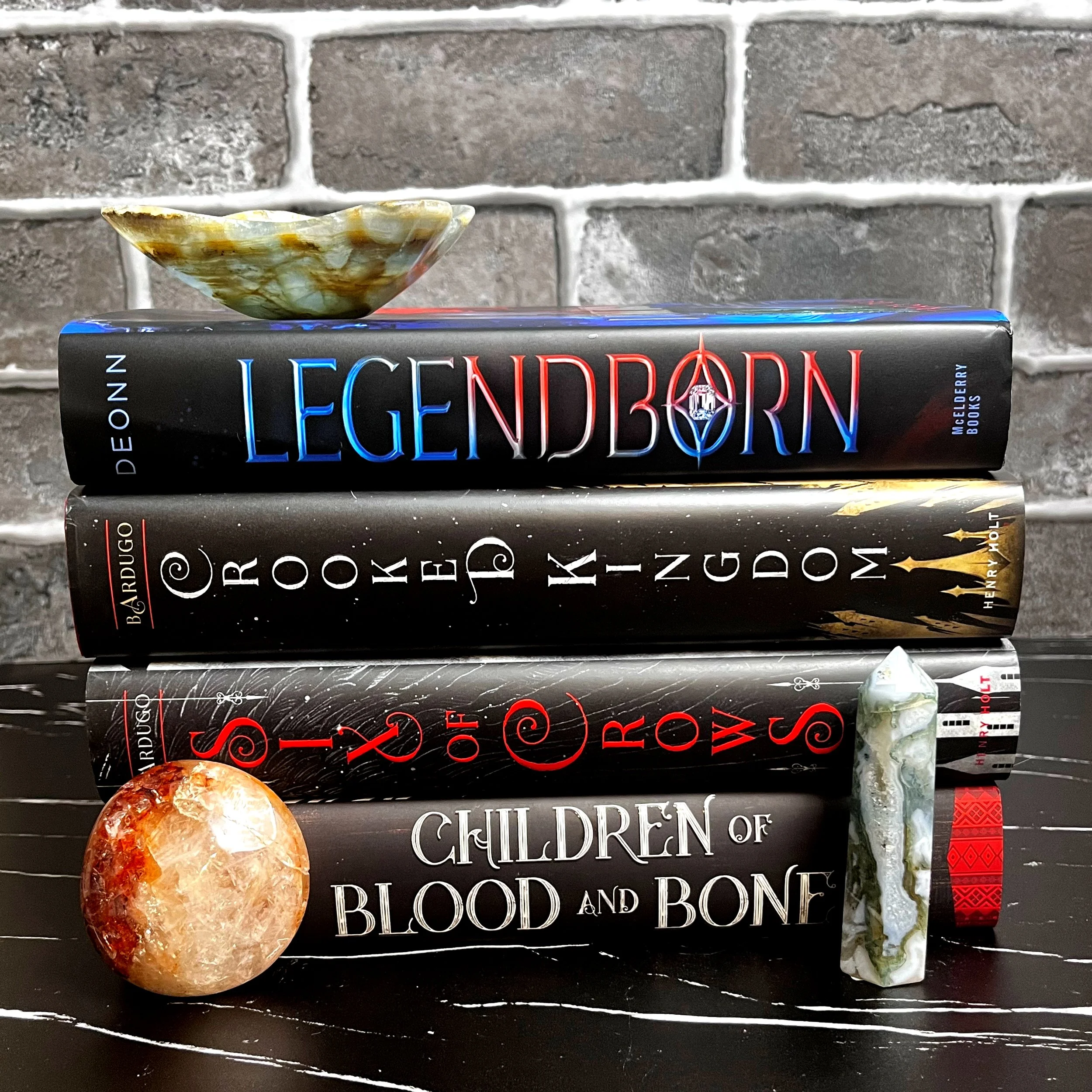 Four black hardcover books stacked in a neat pile with a red palm crystal on the left,  blue and teal tower on right, and a blue green bowl on top of books. Set against grey brick background on top of black marble.