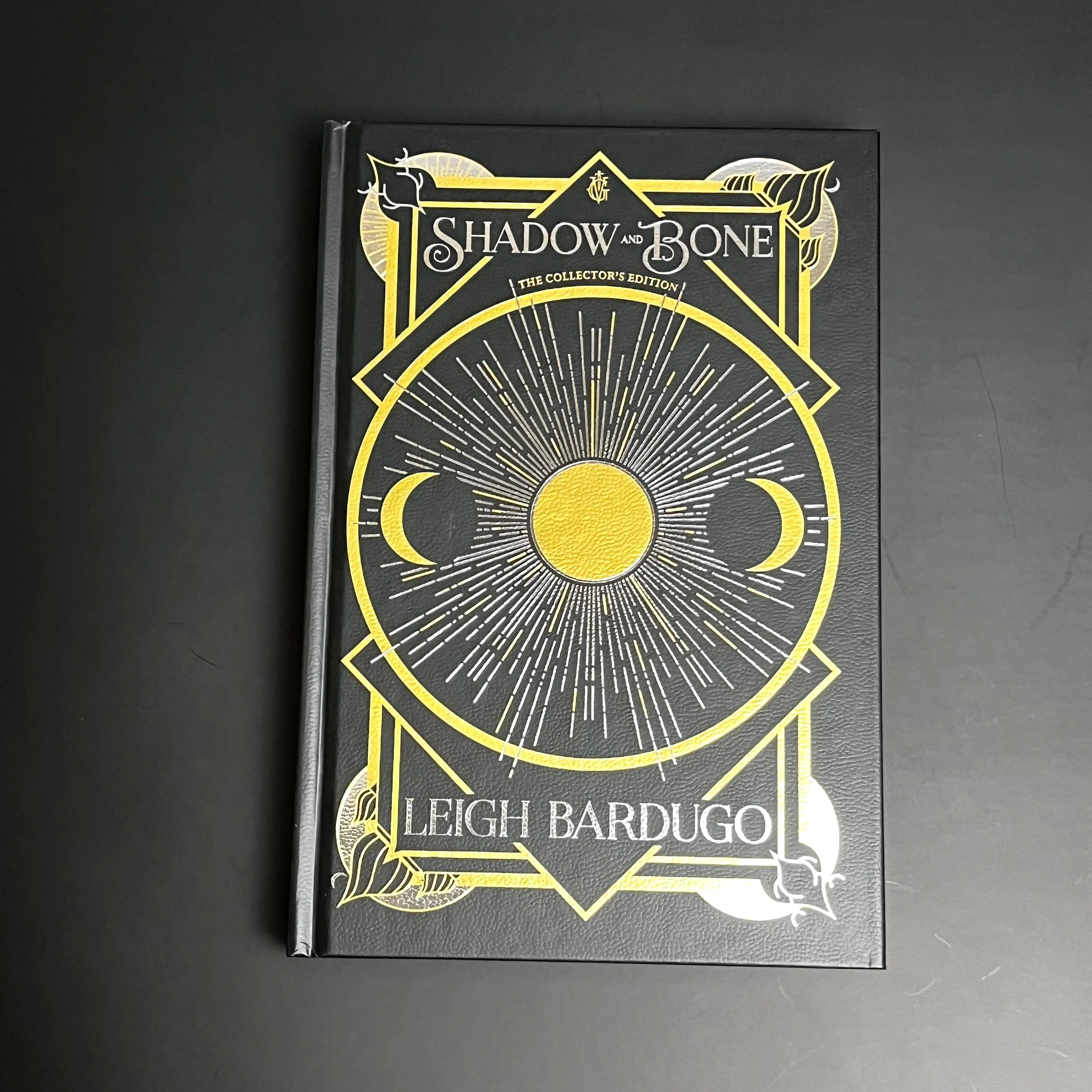Black matte hardcover with leatherlike texture, metallic golden depiction of sun, moon, rays of light within a golden circle and title "Shadow and Bone" at top.