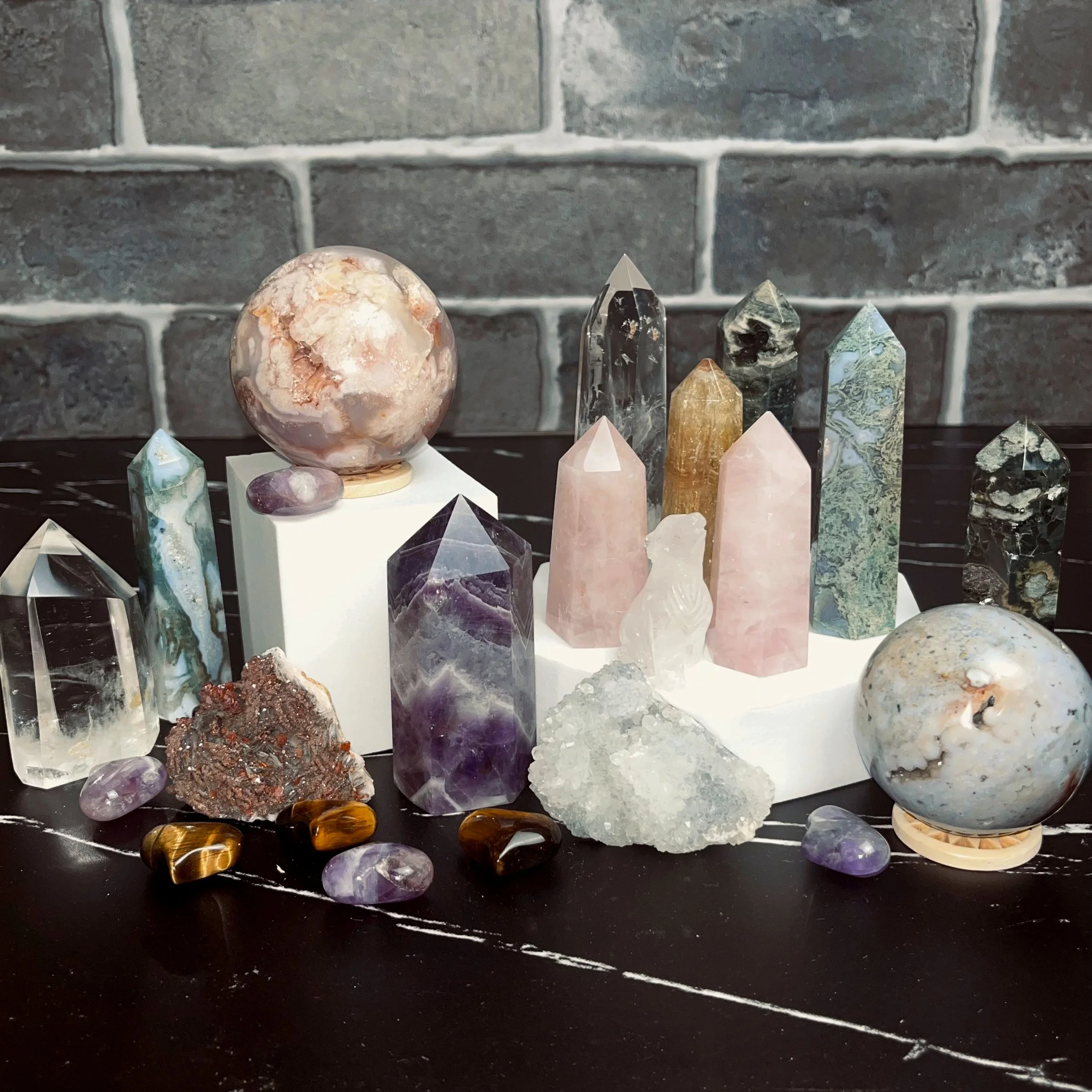 Various crystals, including multiple towers, spheres, and a carved wolf, and two raw Specimen against a black marble counter and a brick background.