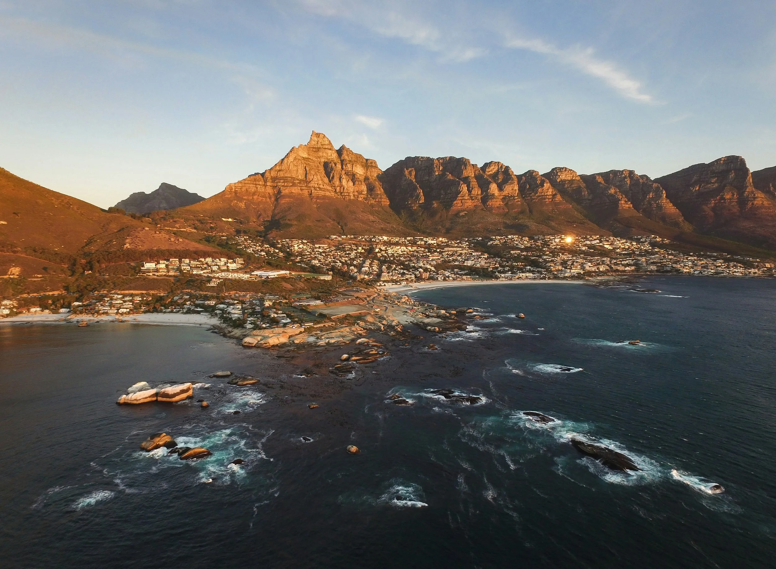 Aerial view of a coastal city at sunset with mountains in the background, ocean waves along the shoreline, and buildings spread across the hillside.