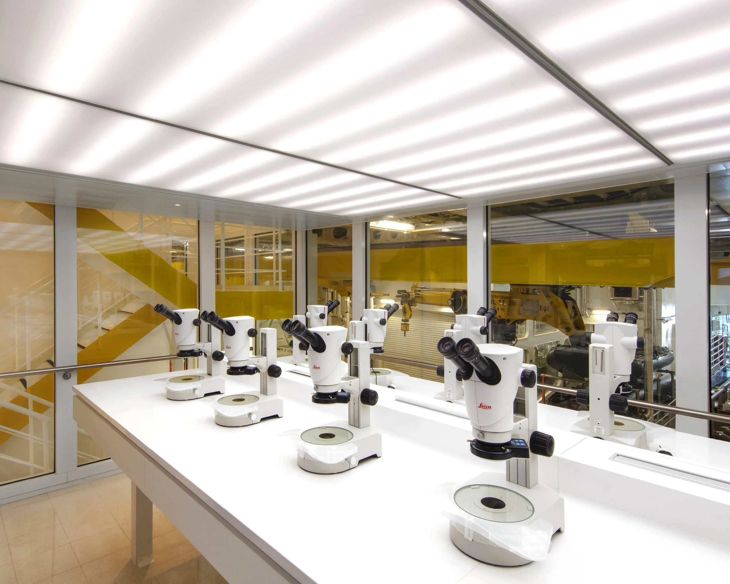 Multiple white microscopes set up on a white table in a modern lab, with large glass windows revealing robotic machinery and yellow structural elements outside.