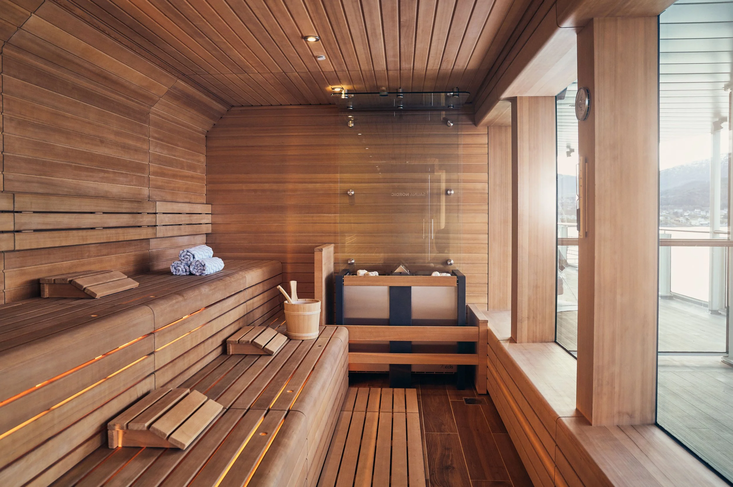A luxurious sauna room with wooden benches, towels, a bucket, and a heater, with large windows letting in natural light and showing a scenic view outside.