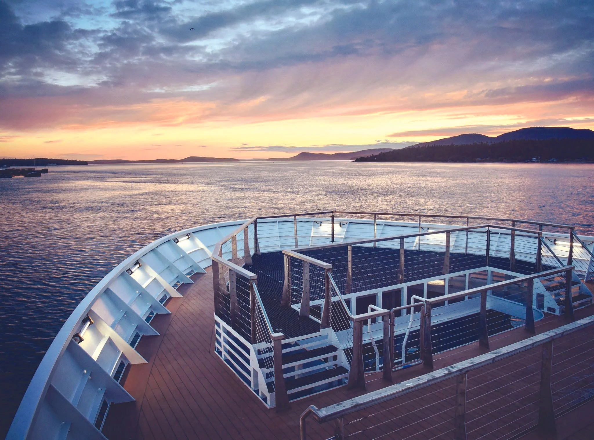 View of a ship's deck with a circular lounge area, railing, and stairs, overlooking a calm body of water during sunset with distant hills and a colorful sky.