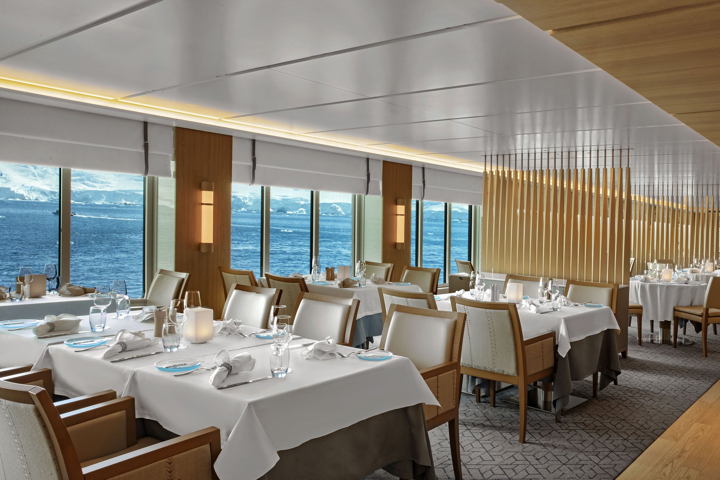 Elegant restaurant with tables set for dining, large windows revealing an ocean view with icebergs, and modern decor with wooden accents.